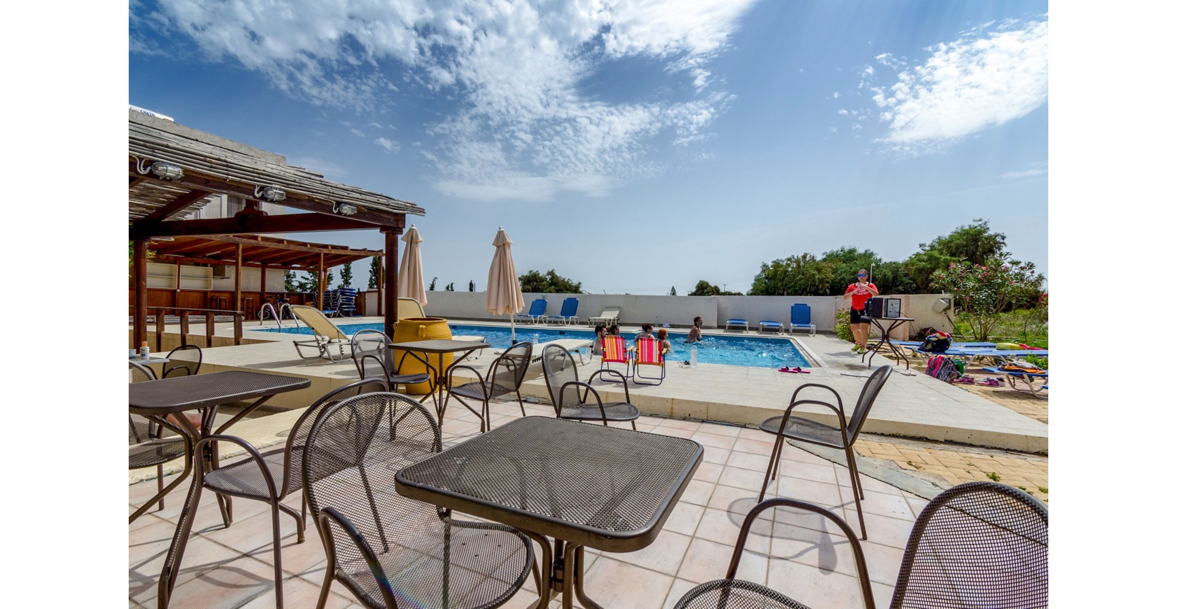Outdoor resort pool area with metal tables and chairs on a tiled terrace. Several people relax and play in the swimming pool under a bright blue sky with scattered clouds. Sun loungers, umbrellas, and a shaded wooden structure frame the poolside setting. The scene conveys a casual, family-friendly vacation atmosphere in warm daylight.