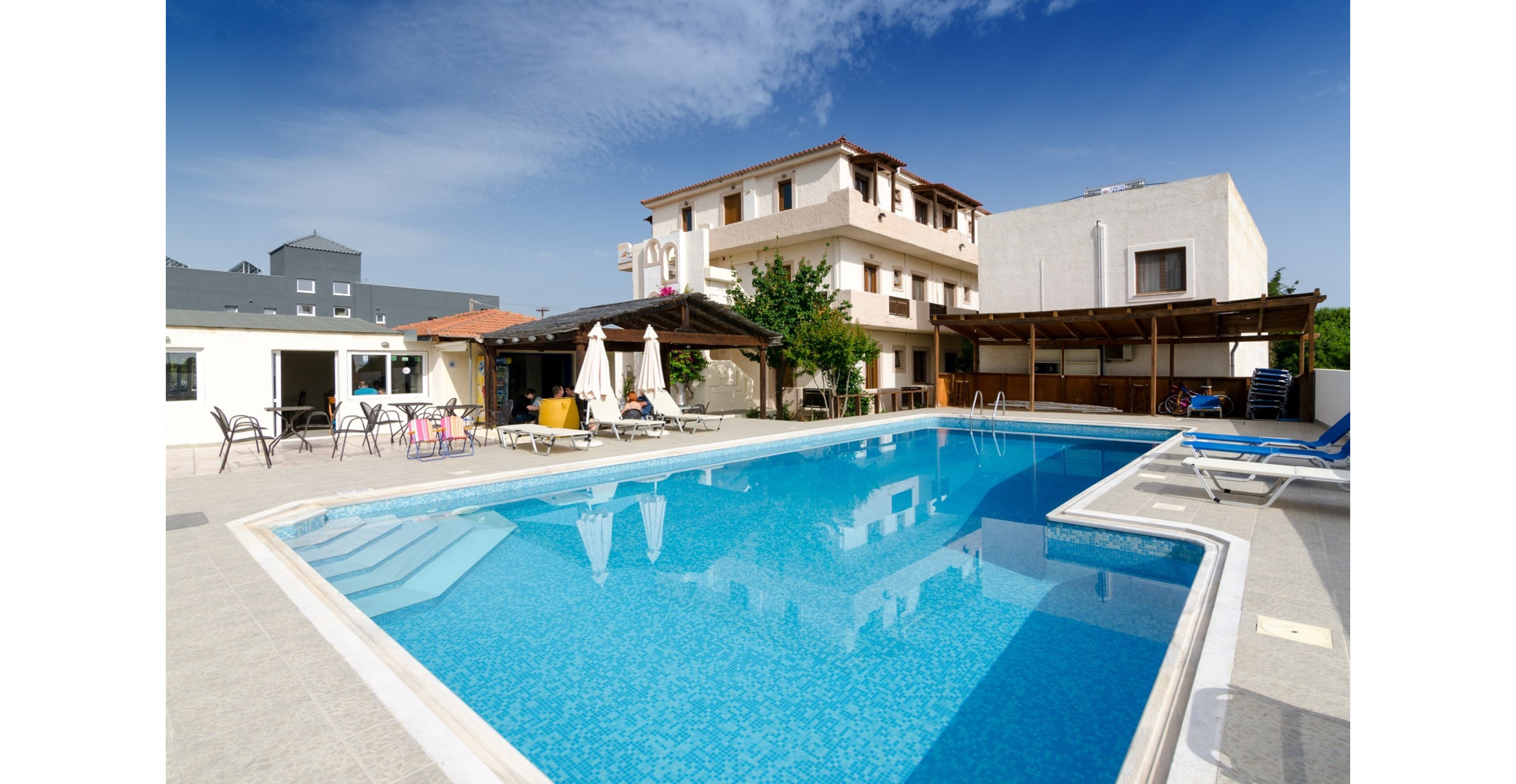 A clear blue swimming pool sits at the center of a small hotel courtyard. Sun loungers and patio chairs line the tiled pool deck under a bright, cloudless sky. Several low-rise buildings with balconies and shaded terraces surround the pool area. The scene suggests a relaxed vacation atmosphere in a warm, sunny climate.