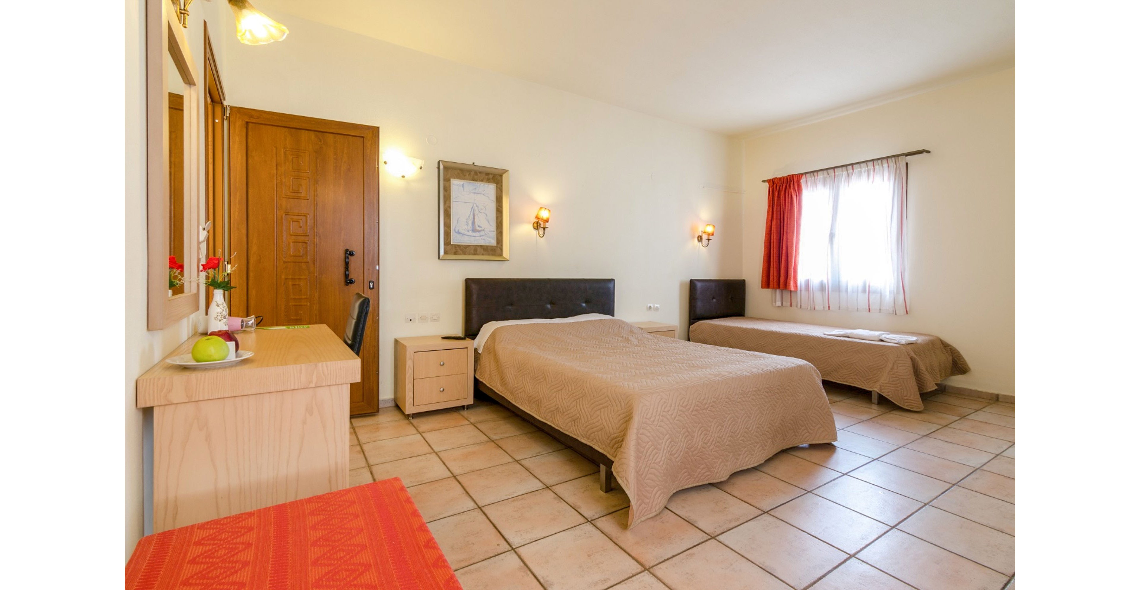 A spacious hotel room features a double bed and a single bed with beige bedspreads. The space includes a wooden door, tiled floor, and a small bedside table between the beds. A desk with a mirror and chair sits to the left, with decorative items and fruit on top. Soft wall sconces and natural light from a curtained window create a warm, inviting atmosphere.
