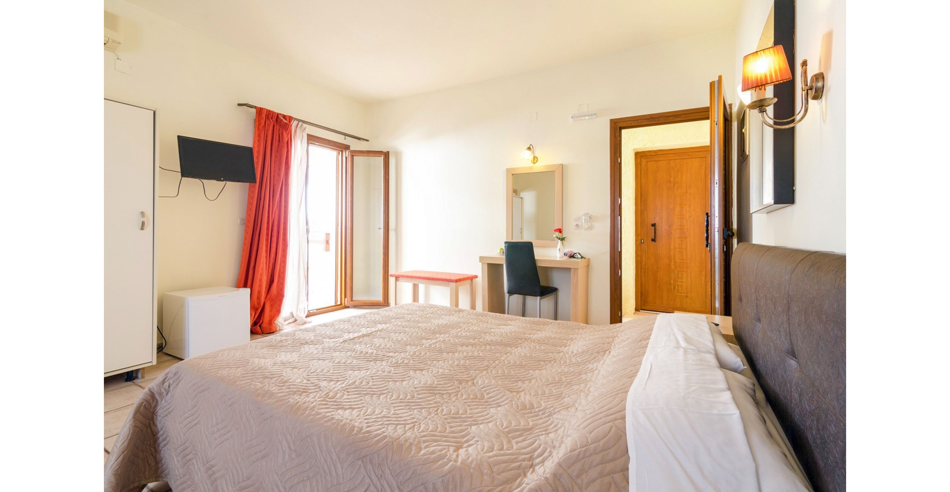 A neatly made double bed dominates a bright hotel-style bedroom. The room features a wooden door, wall-mounted television, and a small desk with a mirror and chair. Sunlight streams in through open glass doors leading to a balcony, with red curtains adding a pop of color. Warm wall sconces and light-colored walls create a welcoming, comfortable atmosphere.