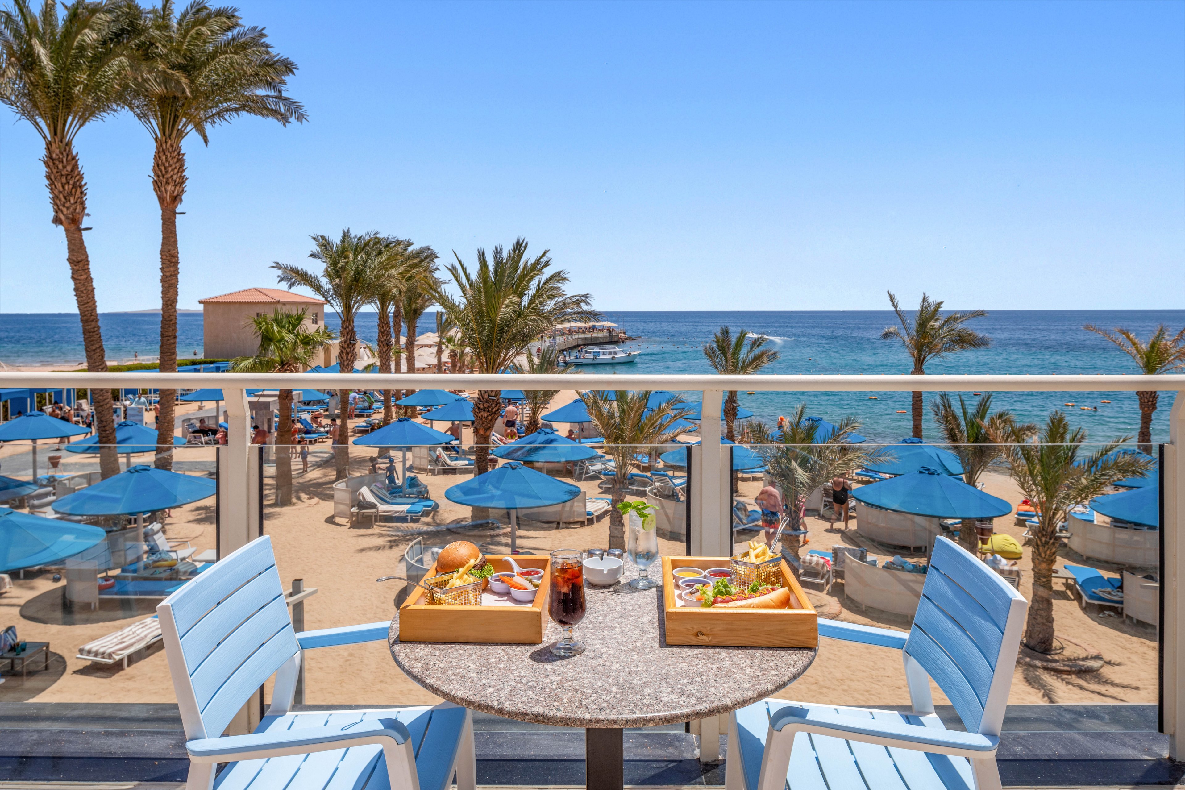 A small round table on a balcony is set with two trays of food and a dark iced drink, framed by two light blue chairs. Beyond the railing, a sandy beach resort is filled with blue sun umbrellas, lounge chairs, and tall palm trees. The clear blue sea stretches to the horizon under a bright cloudless sky, creating a relaxed vacation atmosphere. The scene emphasizes outdoor dining with a panoramic ocean view.