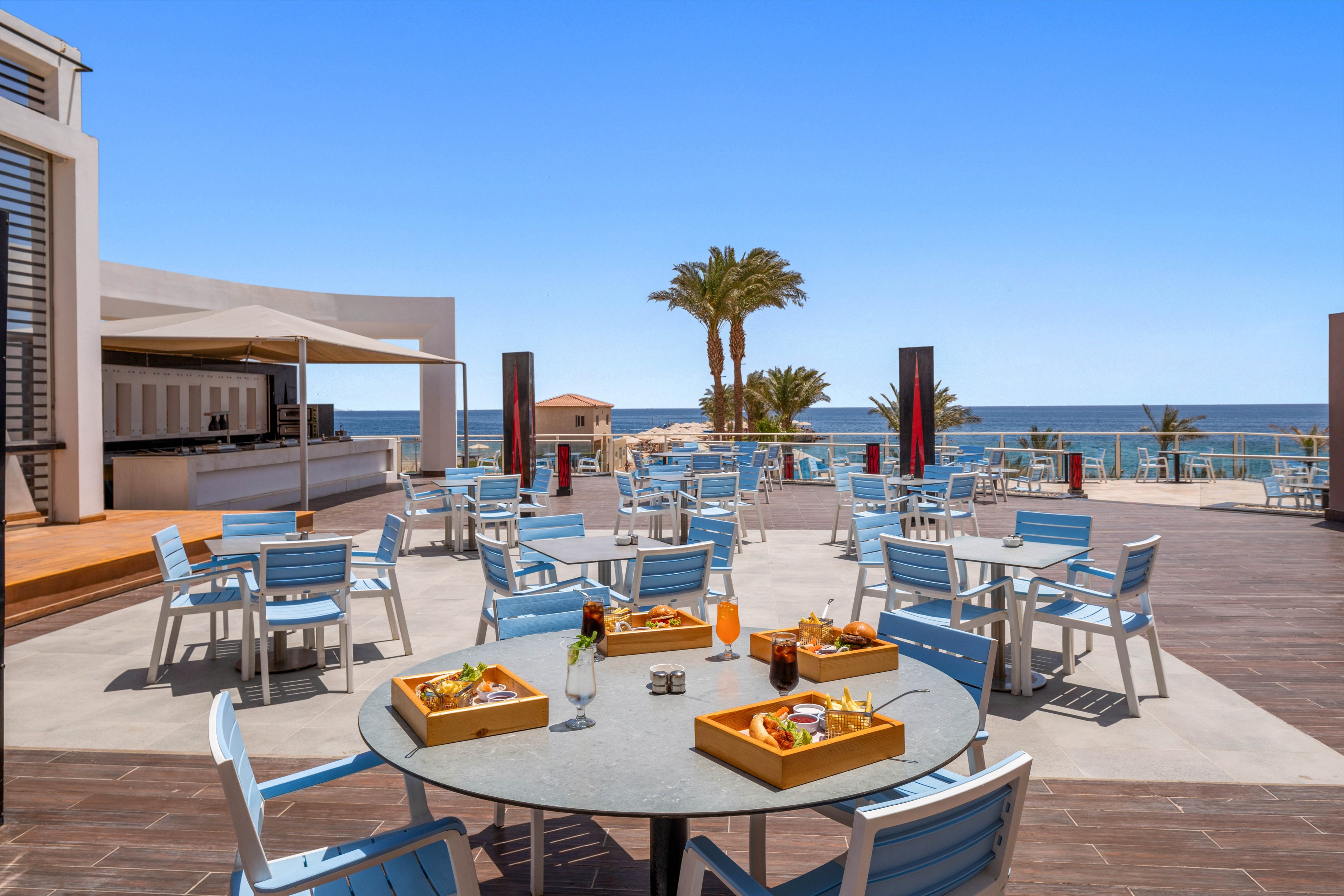 An open-air restaurant terrace features light blue chairs and round tables set with wooden trays of snacks and drinks. The setting overlooks a bright blue sea under a clear sky, with palm trees and resort buildings in the background. A bar counter with shade structure stands to the side, suggesting a relaxed vacation atmosphere. The scene is sunlit and inviting, emphasizing leisure dining by the water.