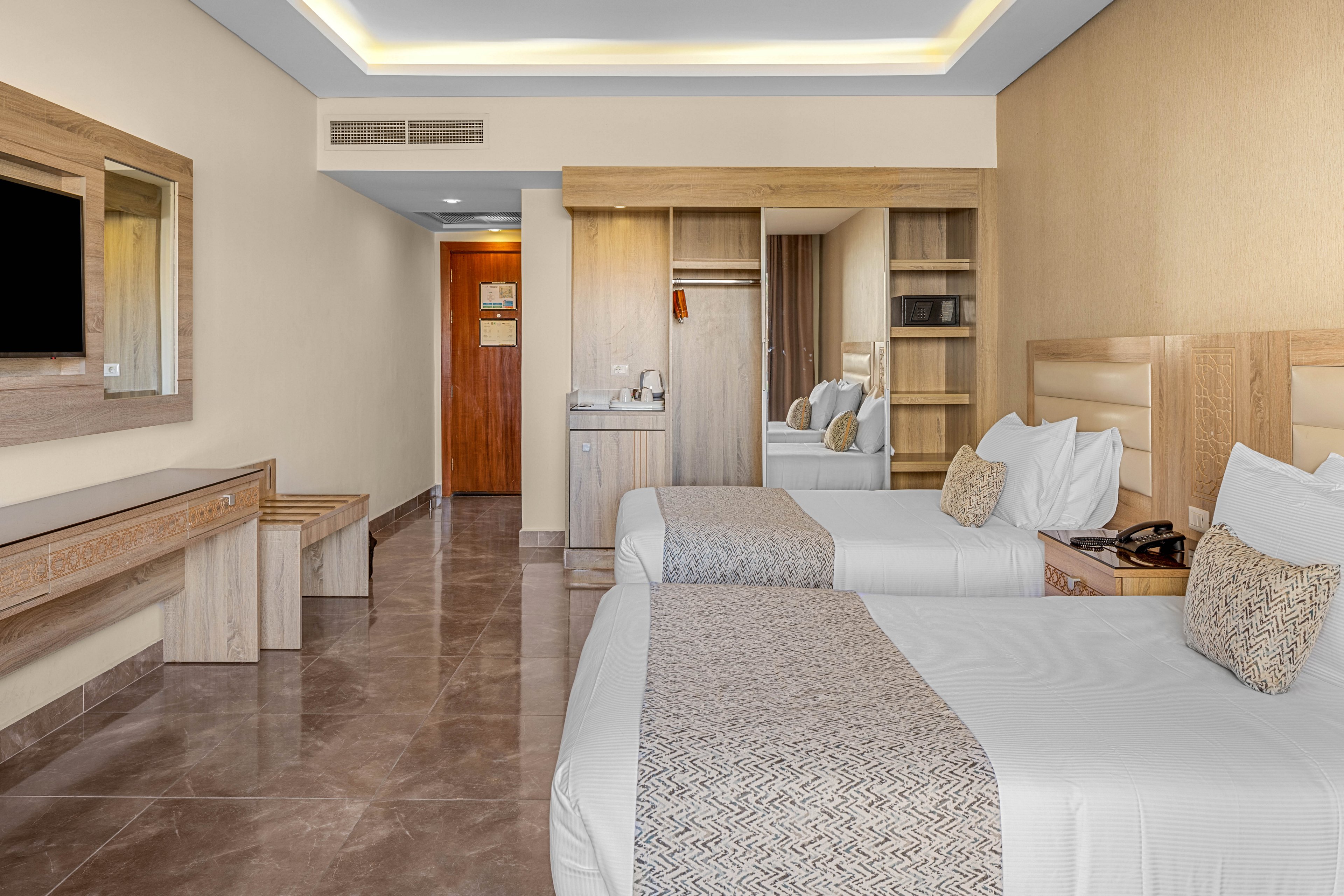 Contemporary hotel room interior featuring two neatly made twin beds with patterned runners and accent pillows. The space includes a wooden desk with mirror, bench seating, and a wardrobe with open shelving. A flat-screen television is mounted on the wall opposite the beds, and a wooden door leads to the hallway. Neutral tones, clean lines, and soft ceiling lighting create a calm, minimalist atmosphere suitable for travelers.