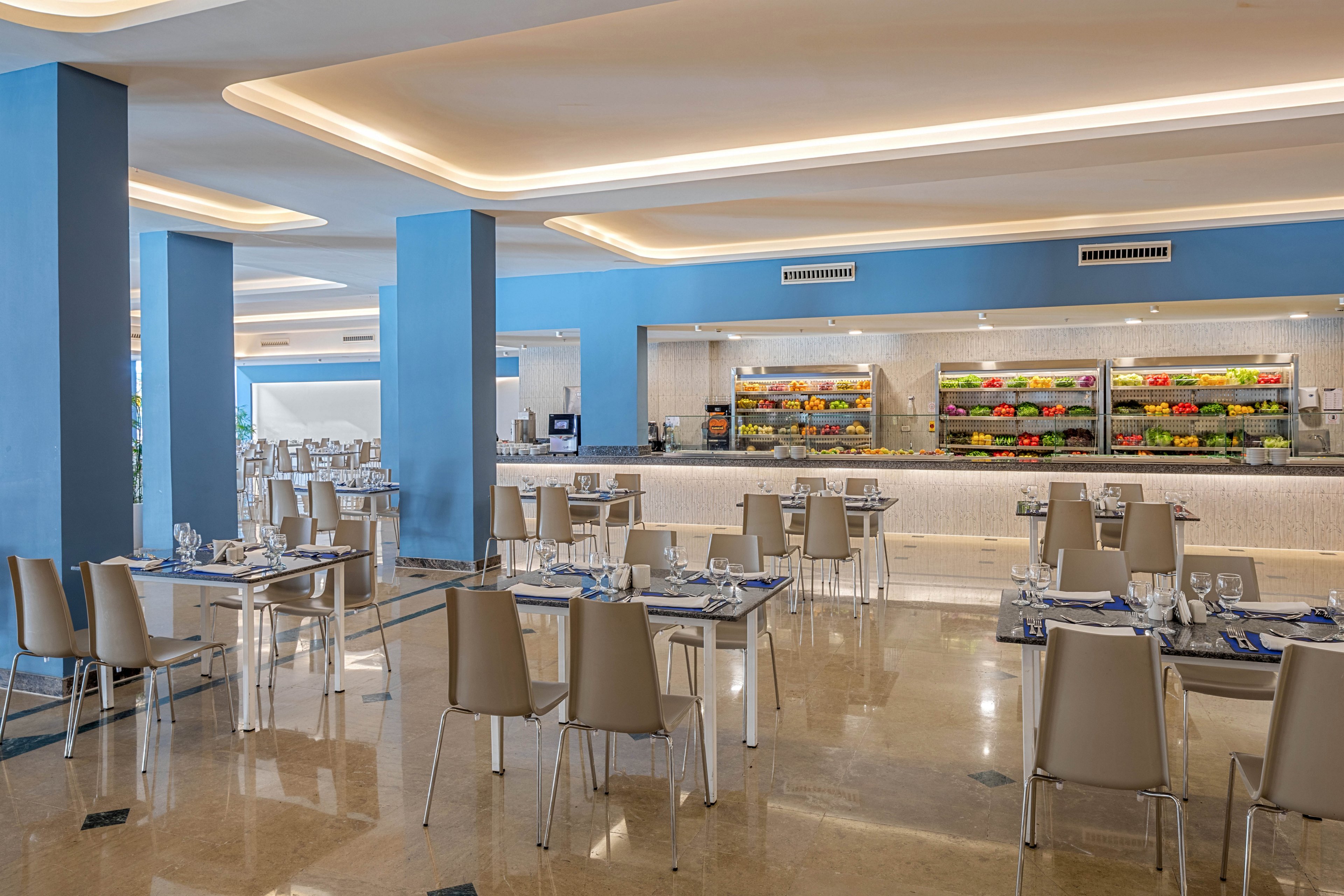 A spacious modern restaurant dining area features neatly arranged tables and beige chairs. The setting appears to be a hotel or resort with polished floors and blue accent walls. In the background, a brightly lit buffet counter displays rows of colorful fruits and vegetables. The atmosphere is clean, organized, and inviting, with table settings prepared for guests.