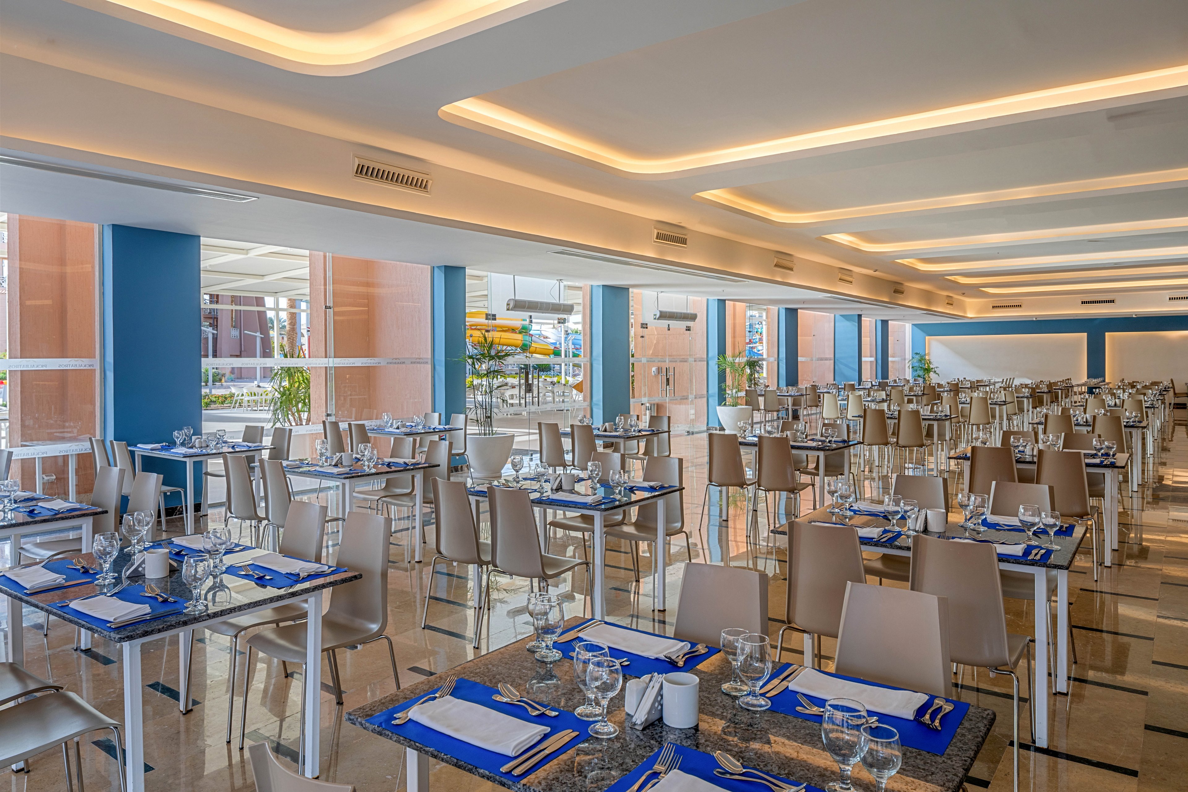 A spacious modern restaurant dining area is shown with rows of neatly arranged tables and chairs. Each table is set with blue placemats, white napkins, cutlery, and glassware, ready for guests. Large windows and glass panels allow natural light to brighten the interior, complemented by warm recessed ceiling lighting. The overall atmosphere feels clean, organized, and contemporary, suggesting a hotel or resort setting.