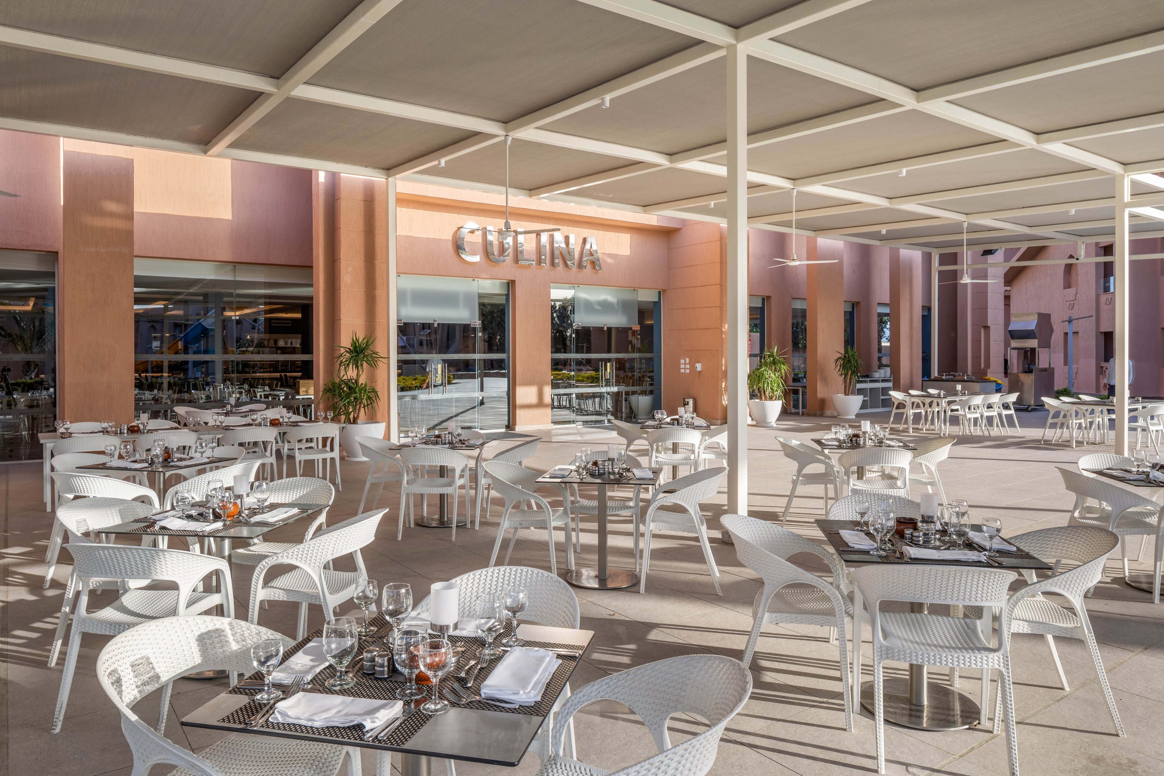 An elegant outdoor restaurant terrace features neatly arranged white tables and chairs under a large canopy. Place settings with glassware, cutlery, and napkins are prepared for guests, suggesting a ready-to-serve dining experience. The background shows a peach-colored building facade with large windows and the word "CULINA" prominently displayed above an entrance. Soft natural light enhances the clean, modern aesthetic of the space.