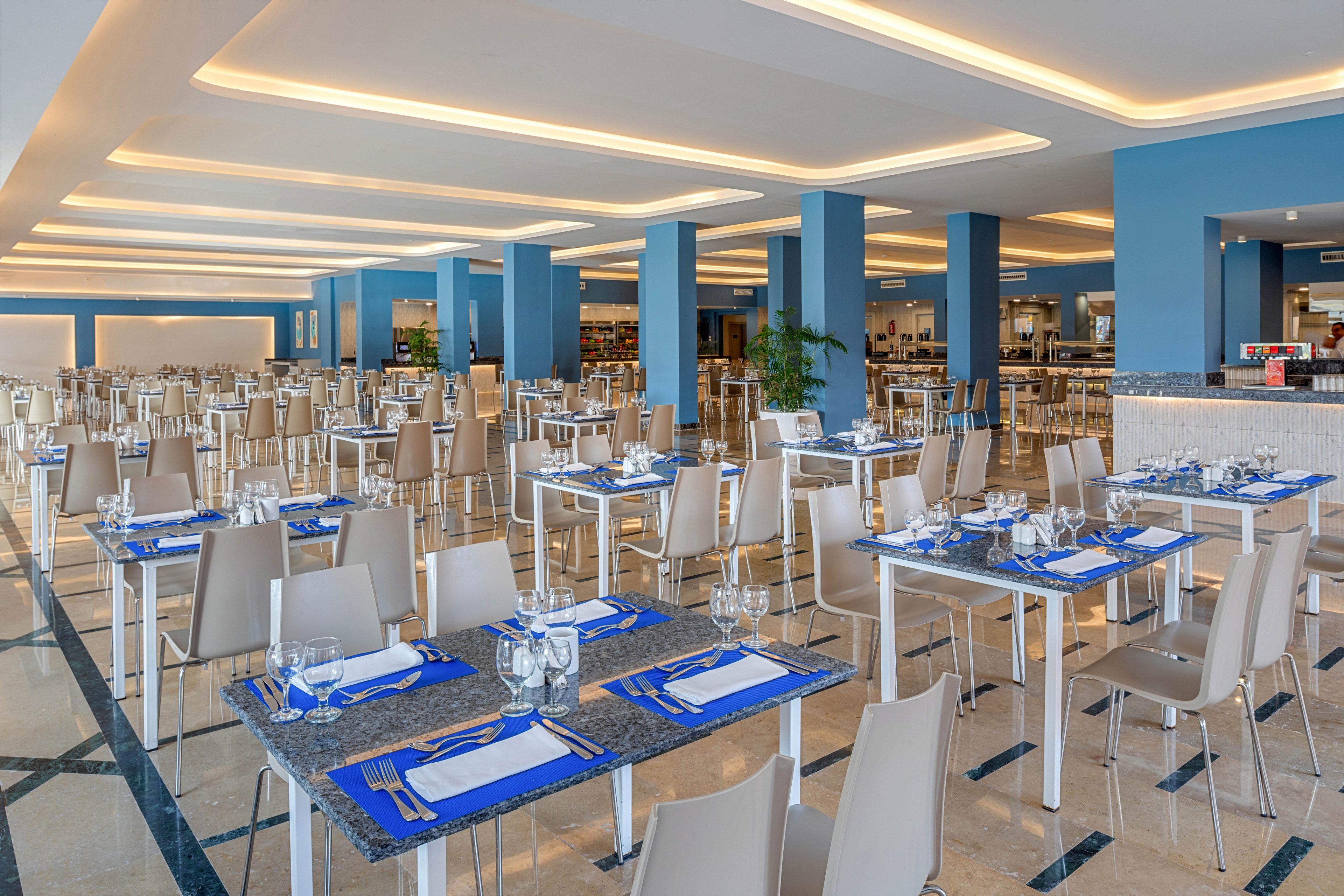 A spacious, modern restaurant dining area is shown fully set and ready for guests. Rows of light-colored tables and chairs are neatly arranged, each table prepared with blue placemats, white napkins, glassware, and cutlery. The interior features blue columns, a polished floor, and a bright ceiling with recessed lighting that creates a warm, inviting glow. In the background, buffet counters and additional seating suggest a hotel or resort dining setting.
