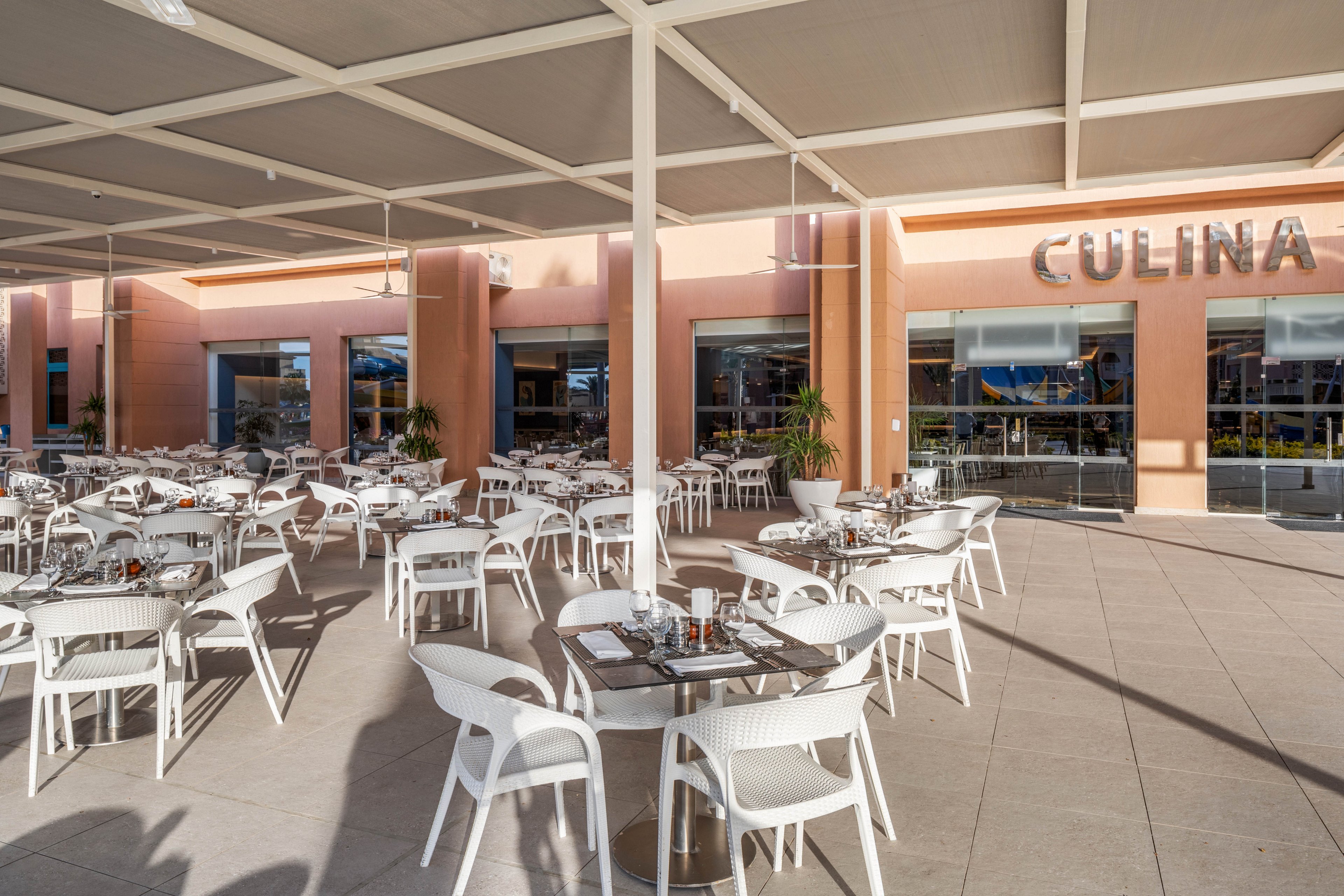 An expansive outdoor restaurant terrace features numerous white tables and chairs neatly arranged under a large canopy. The setting appears calm and ready for service, with place settings and condiments already on the tables. A peach-colored building forms the backdrop, with large glass doors reflecting the bright daylight. The word "CULINA" is prominently displayed above one of the entrances, indicating the venue name.