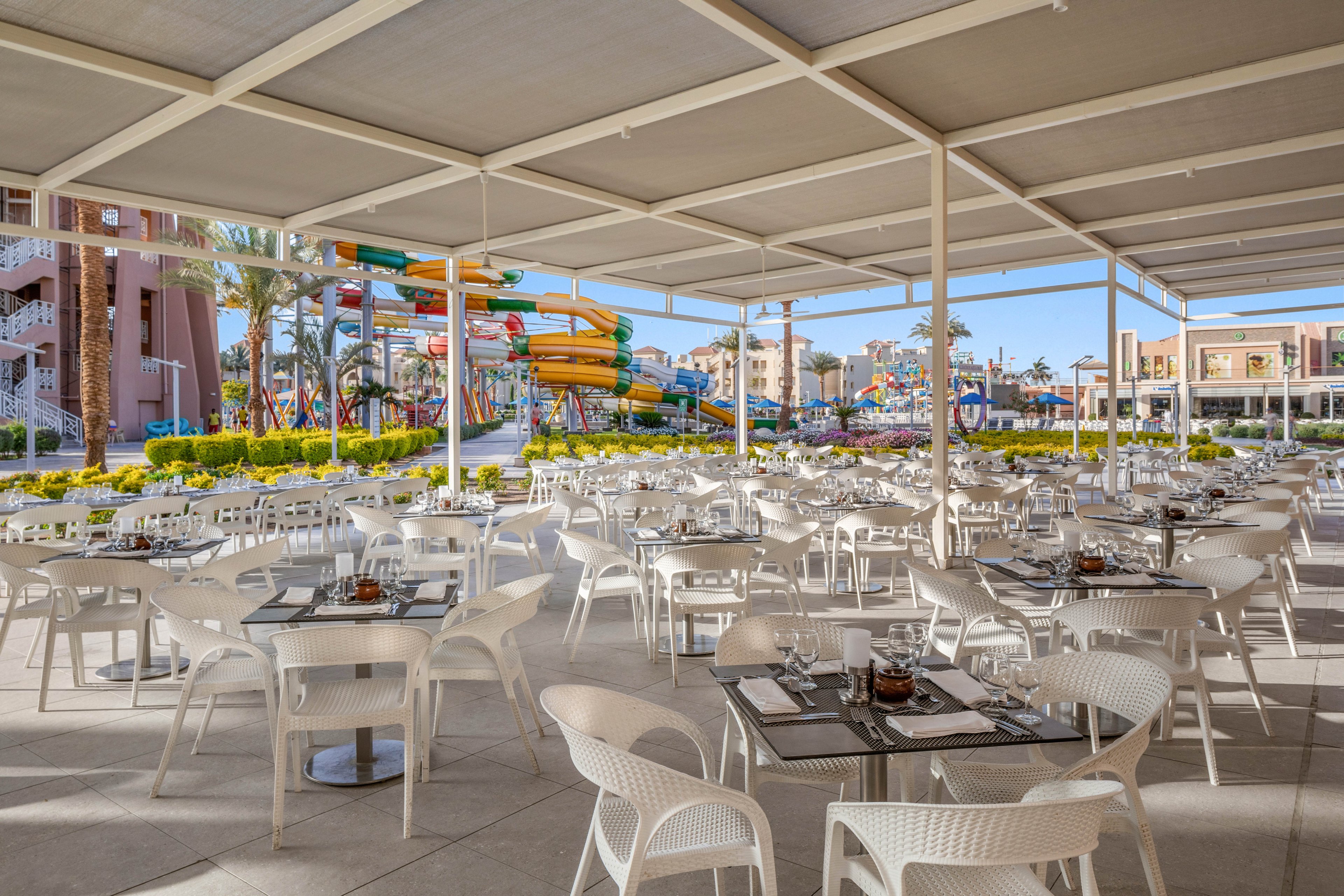 An open-air restaurant with numerous white tables and chairs sits under a large shaded pergola. The dining area is neatly set with glass tabletops, cutlery, and condiments, ready for guests. In the background, a vibrant waterpark with multicolored slides and structures adds a playful backdrop. Palm trees and landscaped greenery enhance the relaxed resort atmosphere under clear daylight.