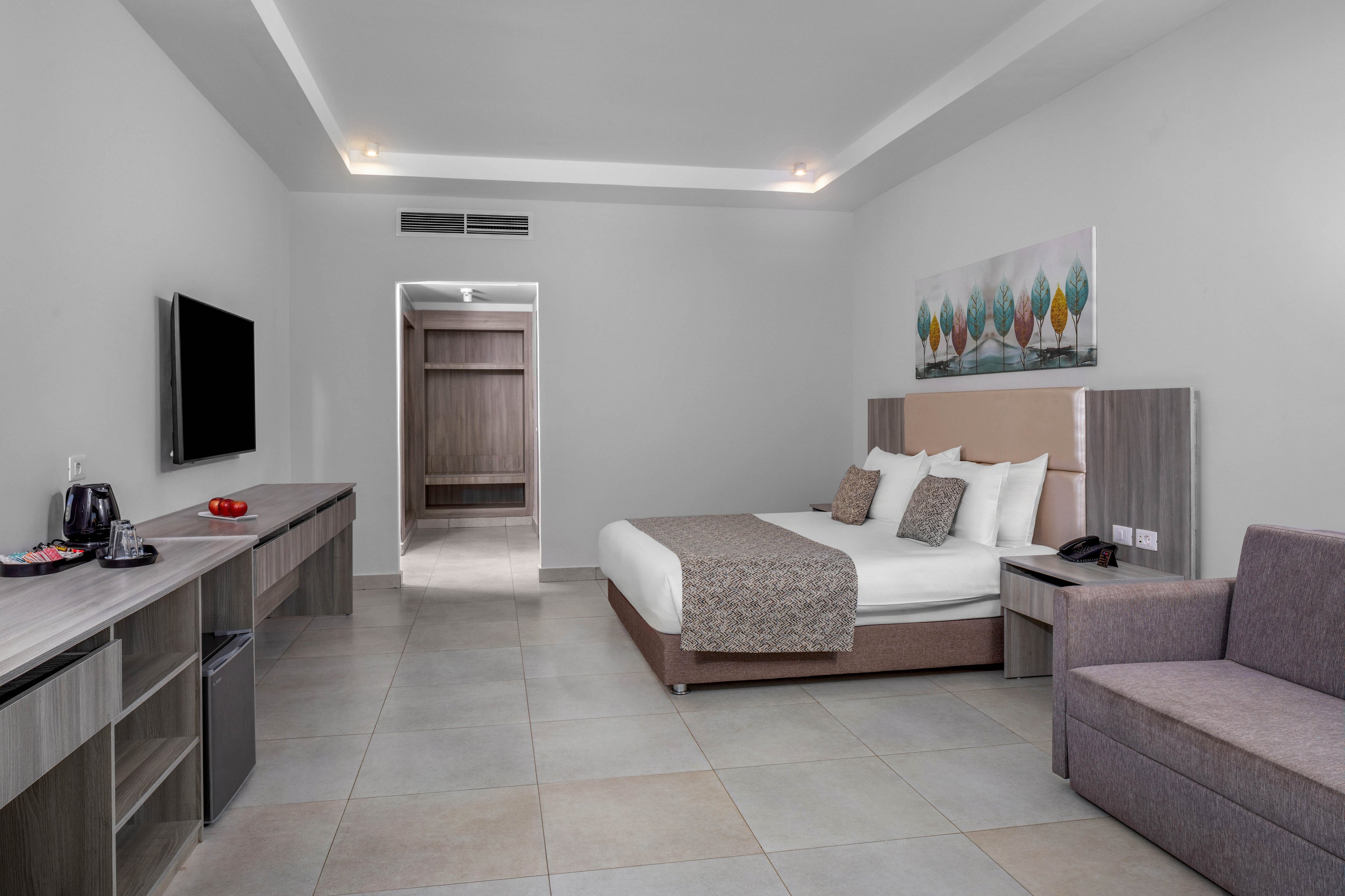 A contemporary hotel room features a large bed with neutral bedding and multiple pillows against a padded headboard. The space includes a wall-mounted flat-screen television, a long desk with storage, and a small seating area with a gray sofa. Soft gray and beige tones dominate the room, complemented by a colorful abstract artwork above the bed. Recessed ceiling lights and an open doorway to a closet or hallway complete the clean, minimalist aesthetic.