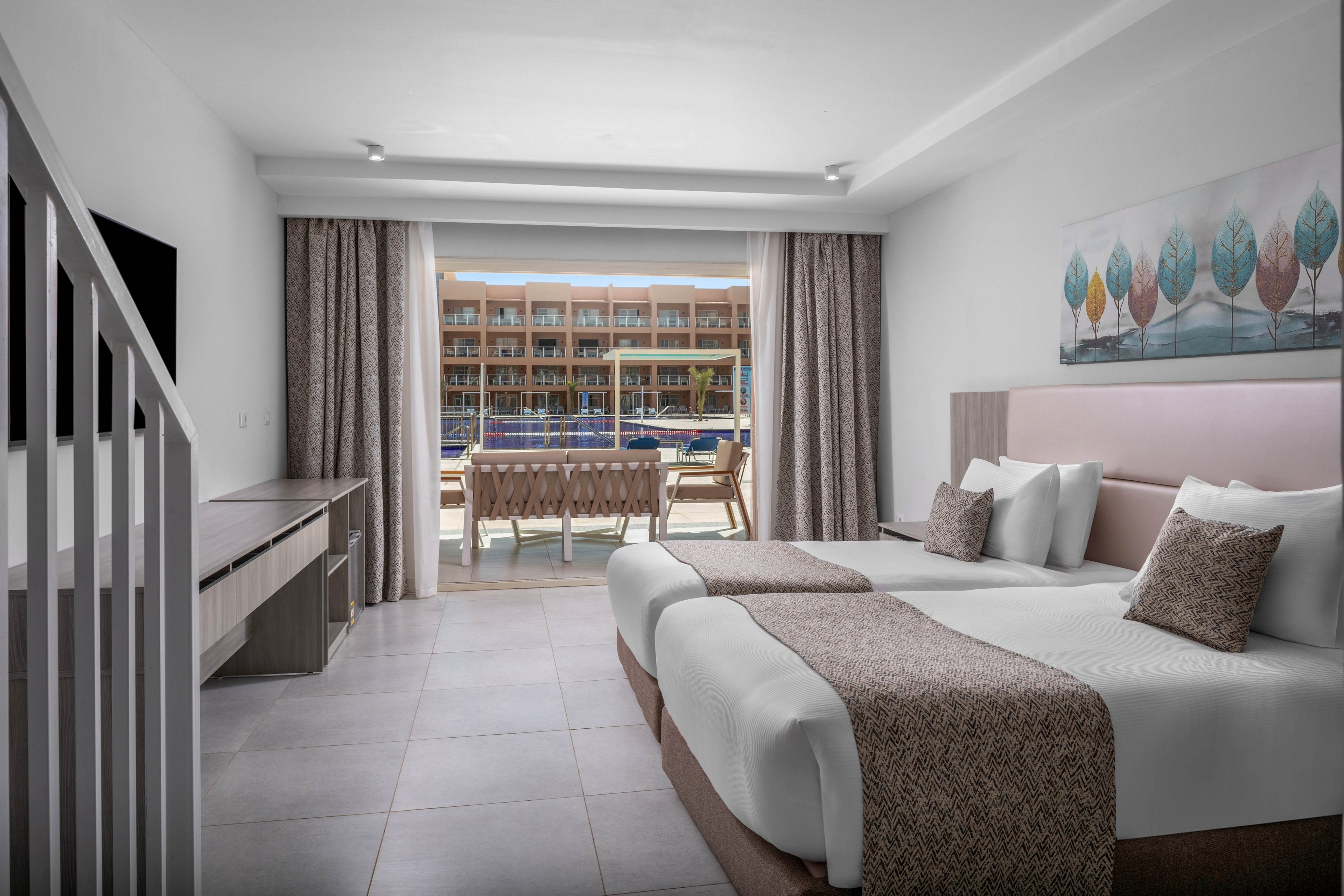 A contemporary hotel room features two neatly made beds with neutral-toned bedding. The space includes a long desk, wall-mounted television, and minimalist decor with a colorful tree artwork above the headboard. Large sliding glass doors open to a balcony with chairs and a table, offering a view of an outdoor pool and adjacent building. Soft natural light and muted colors create a calm, inviting atmosphere suitable for vacation or business travel.