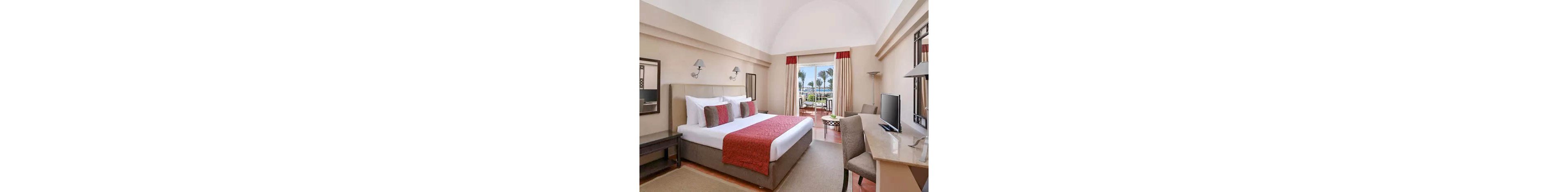 A spacious hotel bedroom features a large bed with white linens and red accents. The room includes bedside lamps, a desk with a television, and neutral-toned furniture. Floor-to-ceiling curtains frame glass doors that open to a balcony with outdoor seating. Palm trees and a distant water view create a relaxing vacation atmosphere.