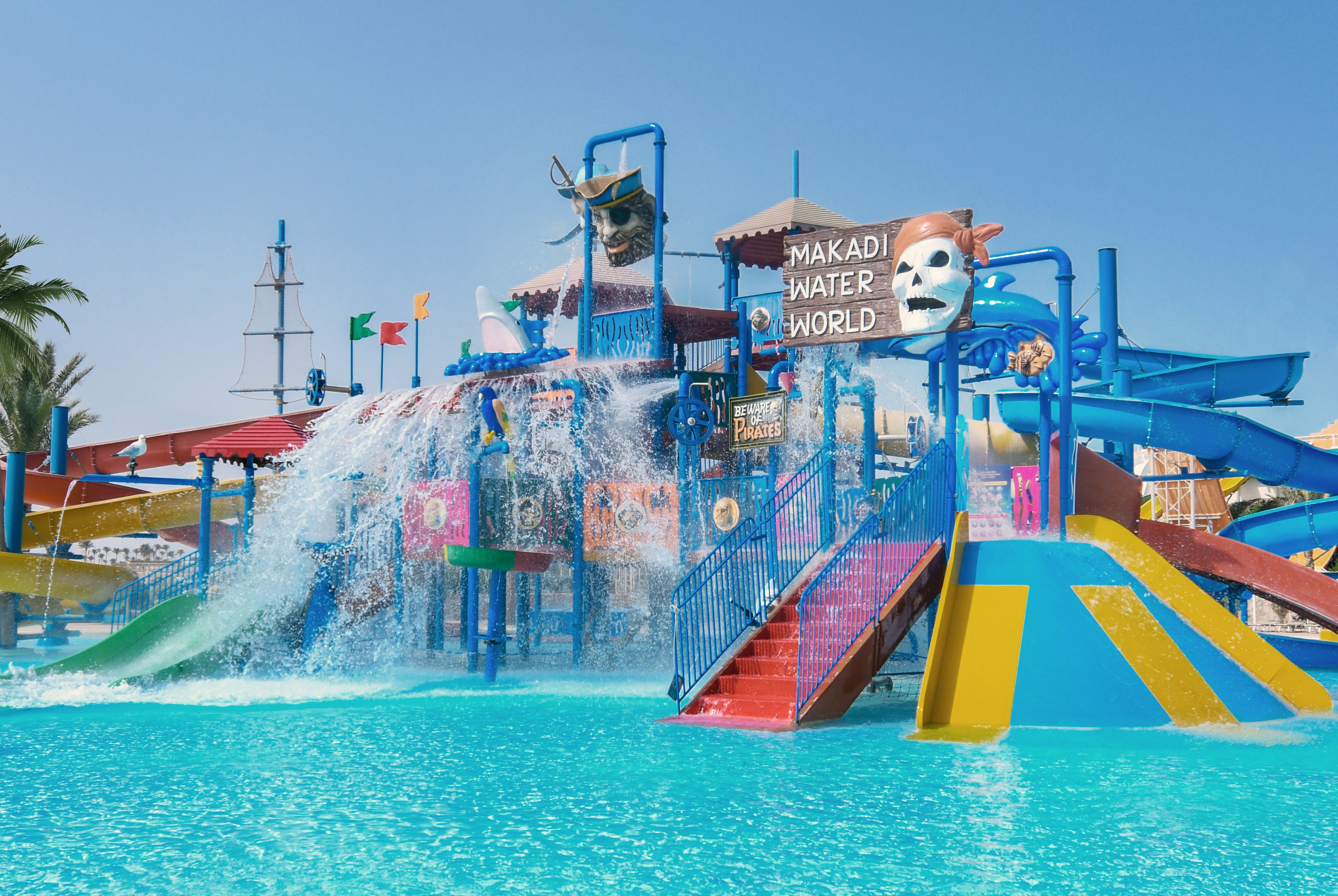 A vibrant water park structure features multiple slides, climbing platforms, and a large tipping bucket splashing water into a bright blue pool. The sign reading "Makadi Water World" is prominently displayed above the attractions. Colorful flags, pirate-themed decorations, and a theatrical mask add playful visual elements. The scene is set under a clear blue sky, emphasizing a fun, family-friendly vacation atmosphere.