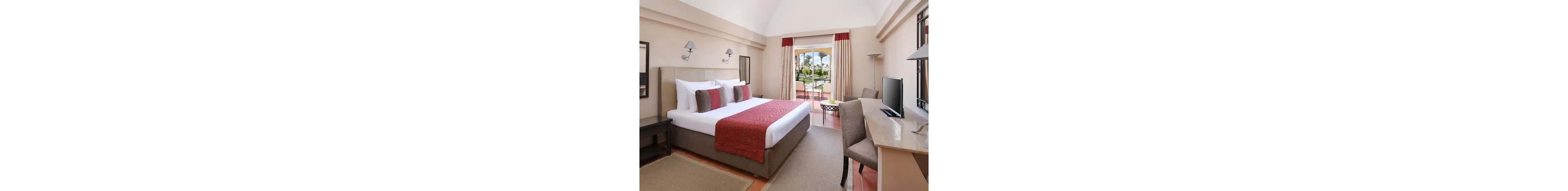A spacious hotel bedroom features a large bed with white linens and red accent pillows and runner. Neutral walls, matching bedside tables, and wall-mounted lamps create a clean, modern look. A desk with a flat-screen television sits opposite the bed, alongside a simple chair. Floor-to-ceiling curtains frame glass doors that open to a terrace overlooking a lush garden with palm trees.