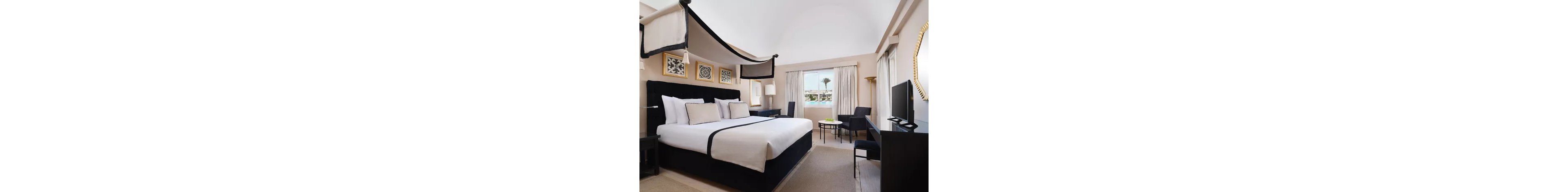 A contemporary hotel room features a large canopy bed with white linens and dark trim. The space includes bedside tables, framed wall art, and a seating area with chairs and a round table. Natural light enters through a window with curtains, revealing a palm tree and bright sky outside. Neutral tones and clean lines create a calm, minimalist atmosphere suitable for relaxation or business travel.