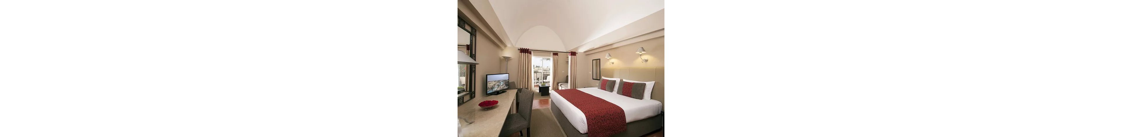 A contemporary hotel bedroom features a large bed with white linens and red patterned accents. A desk with a chair faces a flat-screen television, and decorative mirrors and wall lamps enhance the space. Floor-to-ceiling curtains frame glass doors that open to a balcony with outdoor seating. Neutral walls and soft lighting create a calm, inviting atmosphere suitable for relaxation or business travel.