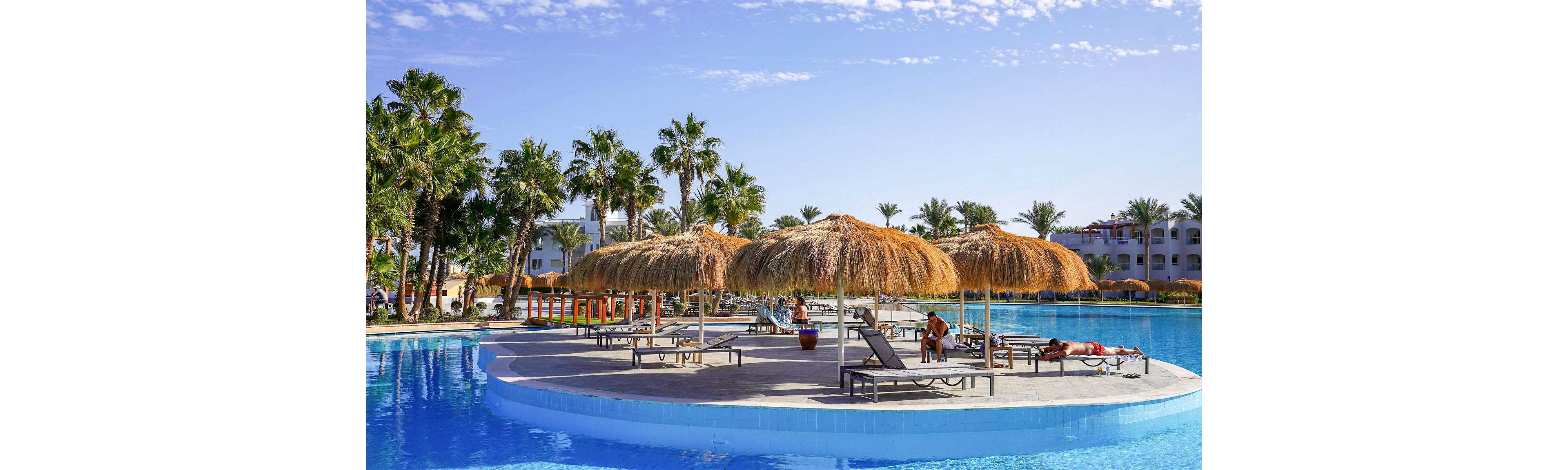 A large resort swimming pool features a circular island deck with straw parasols and sun loungers. People relax on the loungers, sunbathing under clear blue skies. Palm trees surround the pool area, with white hotel buildings visible in the background. The bright sunlight and reflections on the water create a warm, vacation atmosphere.