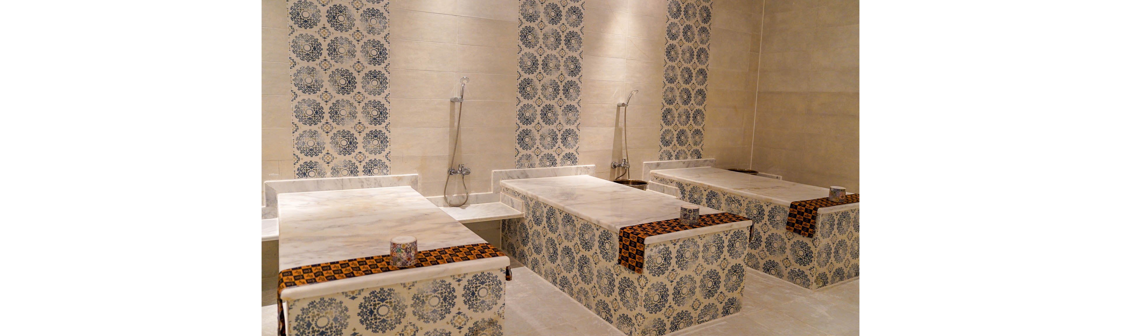 The image shows a traditional hammam interior with multiple marble massage tables arranged in a tiled room. Ornate blue and white patterned tiles decorate the walls and the sides of the tables, creating a classic Middle Eastern aesthetic. Each table has a folded patterned cloth at the head and a small decorative bowl or container placed on top. Wall-mounted handheld shower fixtures are visible, emphasizing the bathing and spa function of the space.