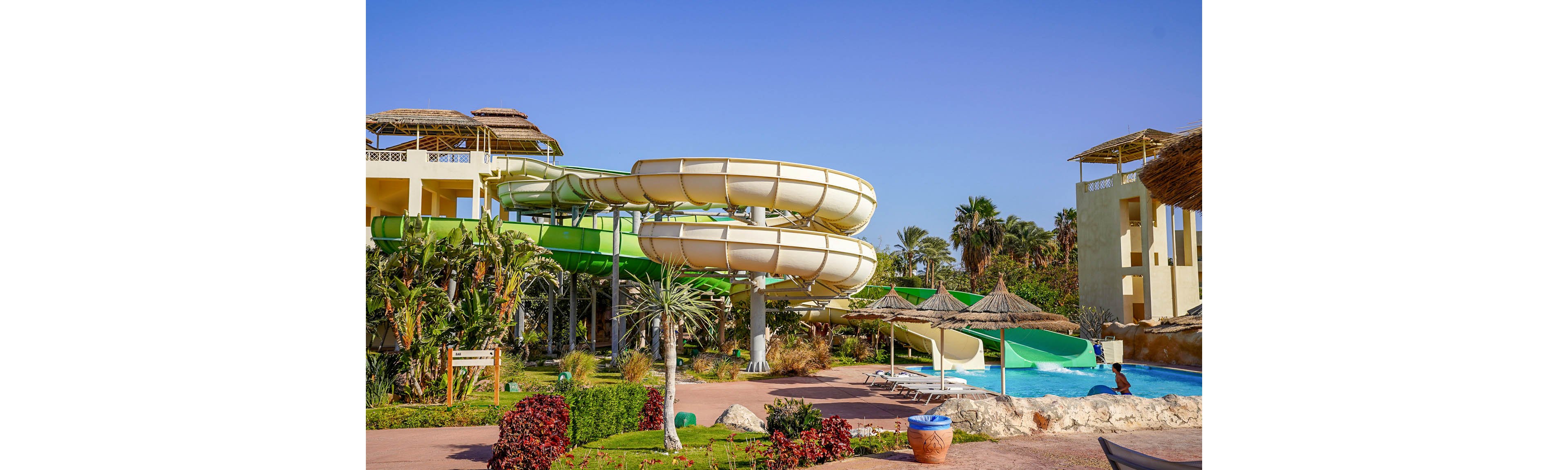 An outdoor resort water park features large spiral water slides in white and green under a clear blue sky. Tropical plants and palm trees surround the slides, creating a lush vacation atmosphere. Thatched sun umbrellas and lounge areas line the edge of a bright blue pool. A person enjoys the water near the slide exit, adding a sense of leisure and fun.