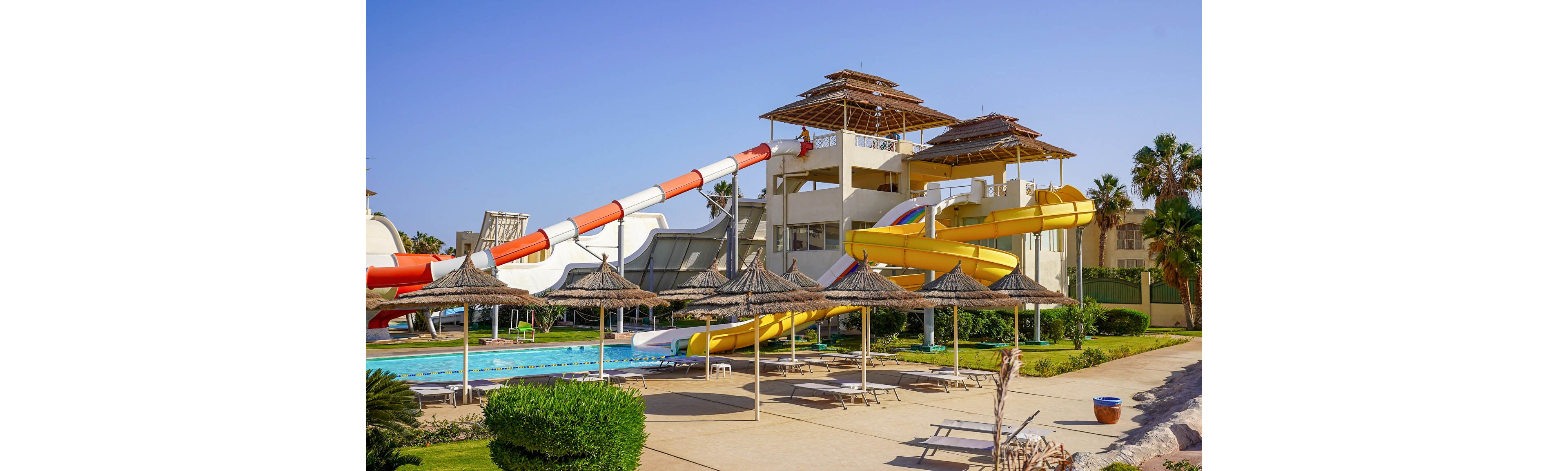 An outdoor resort water park features large twisting slides in bright yellow and red-white colors. The slides descend into a long swimming pool surrounded by sun loungers and straw parasols. A multi-level structure supports the slides, with a person standing near the top platform. Palm trees and manicured lawns add a tropical vacation atmosphere under a clear blue sky.