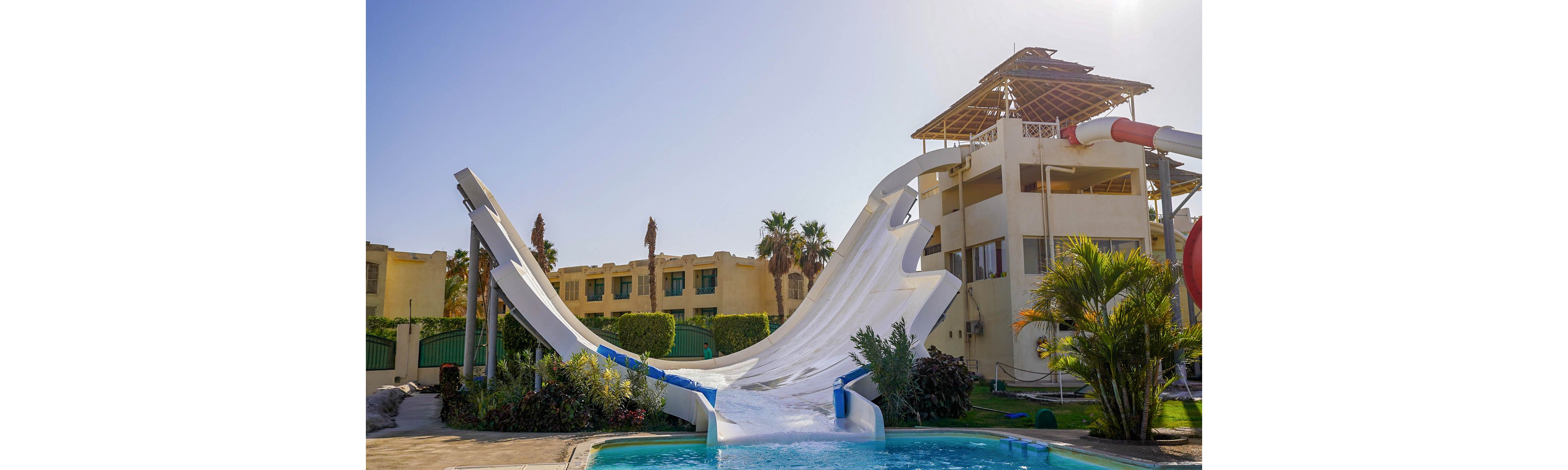 A large white water slide curves dramatically into a clear blue swimming pool at an outdoor resort. The slide structure is attached to a multi-story building with open balconies and a partially covered rooftop. Palm trees, shrubs, and other greenery surround the pool area, adding a tropical feel. Bright sunlight and a clear sky suggest a warm, sunny day at the water park.