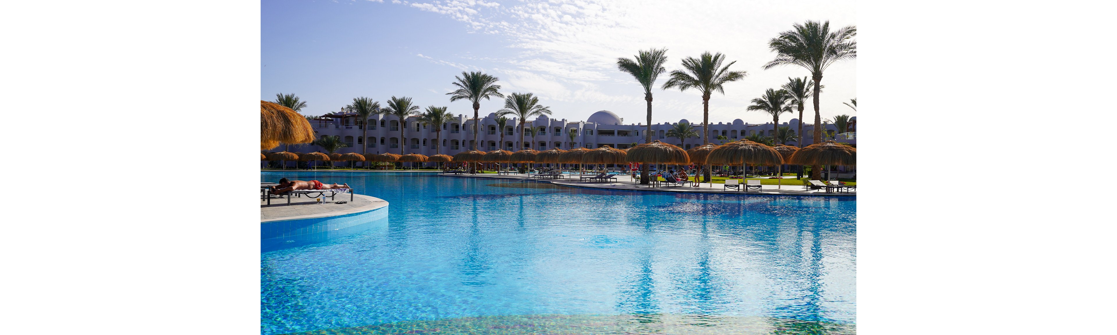 A large, clear blue swimming pool stretches across the foreground of a resort complex. Palm trees and thatched sunshades line the poolside, with lounge chairs arranged for relaxation. White low-rise buildings with domed roofs form the backdrop under a bright, partly cloudy sky. A person is sunbathing on a lounger near the pool, adding a tranquil vacation atmosphere.