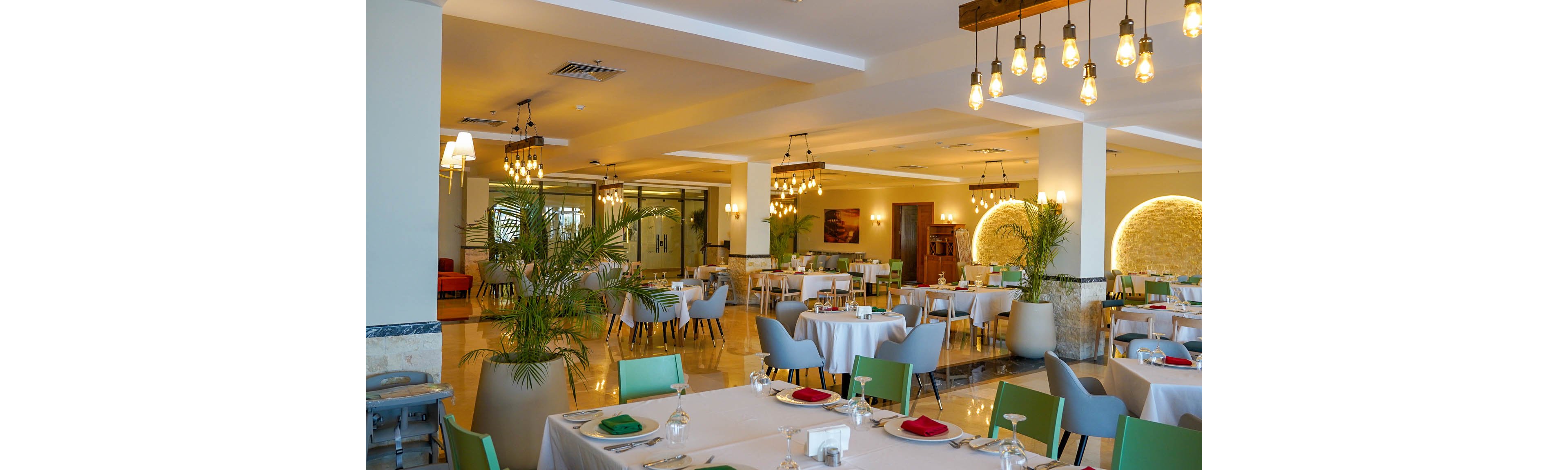 Spacious restaurant dining room with neatly arranged tables and chairs. White tablecloths, glassware, and folded napkins create an elegant yet casual atmosphere. Warm pendant lighting and decorative wall lights illuminate the space, complemented by indoor plants and neutral tones. The room appears ready for guests, with a clean, contemporary design and polished floor surfaces.