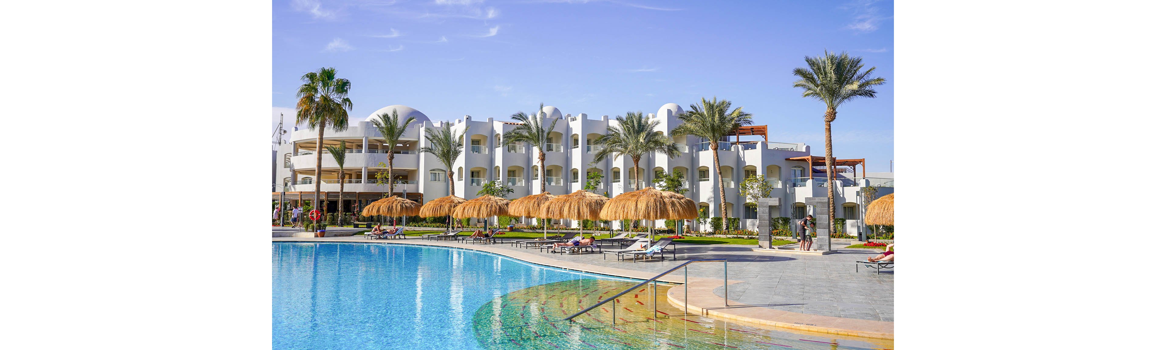 A large outdoor swimming pool sits in front of a white resort hotel with domed roofs. Straw parasols and sun loungers line the poolside, where guests relax in the sun. Tall palm trees and manicured greenery add a tropical atmosphere to the scene. The bright blue sky and clear water create a warm, inviting vacation setting.