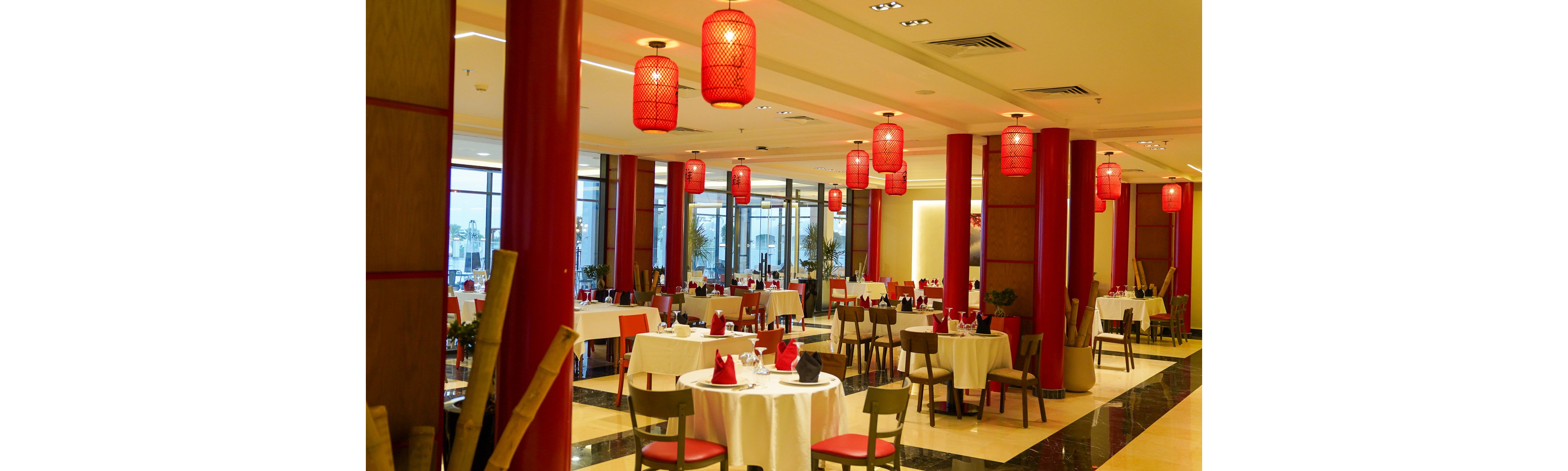 Spacious restaurant dining room featuring round tables with white tablecloths and neatly arranged chairs. The setting includes large red columns and hanging red lanterns that create a warm, inviting atmosphere. Floor-to-ceiling windows line one side of the room, allowing natural light to complement the soft interior lighting. Red accents on napkins and decor enhance the sophisticated, contemporary Asian-inspired style.