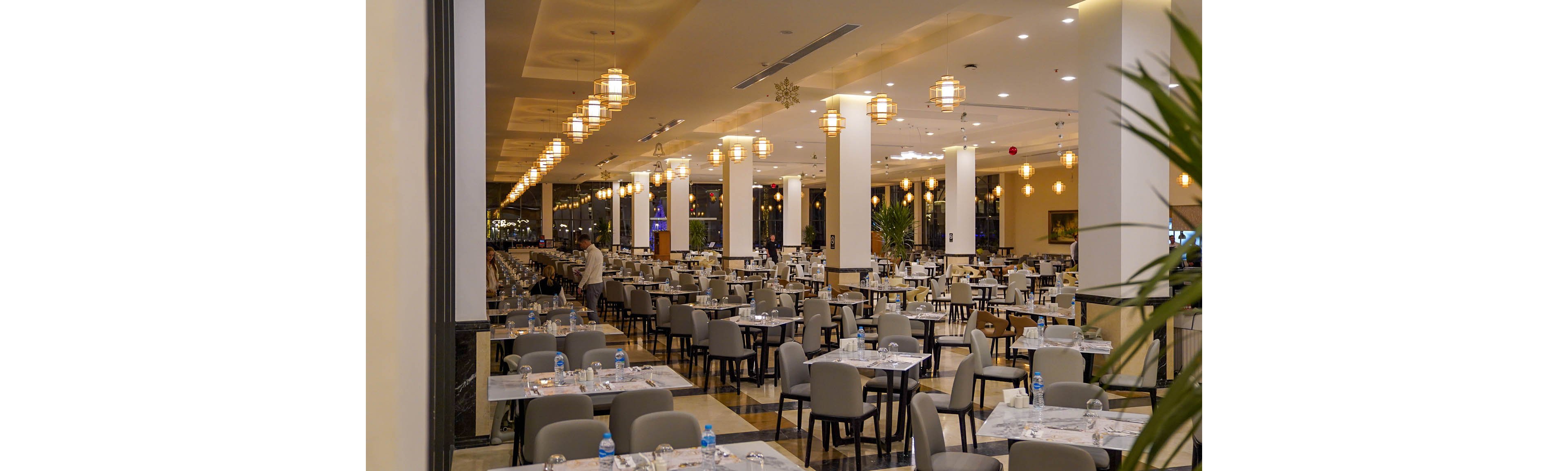 A large, modern restaurant dining hall is filled with neatly arranged tables and gray upholstered chairs. Warm pendant lights hang from the ceiling, creating a cozy atmosphere over the polished floor. Table settings with glassware and bottled water are prepared, suggesting readiness for guests. A few people are visible in the background, adding subtle activity to the otherwise calm interior scene.