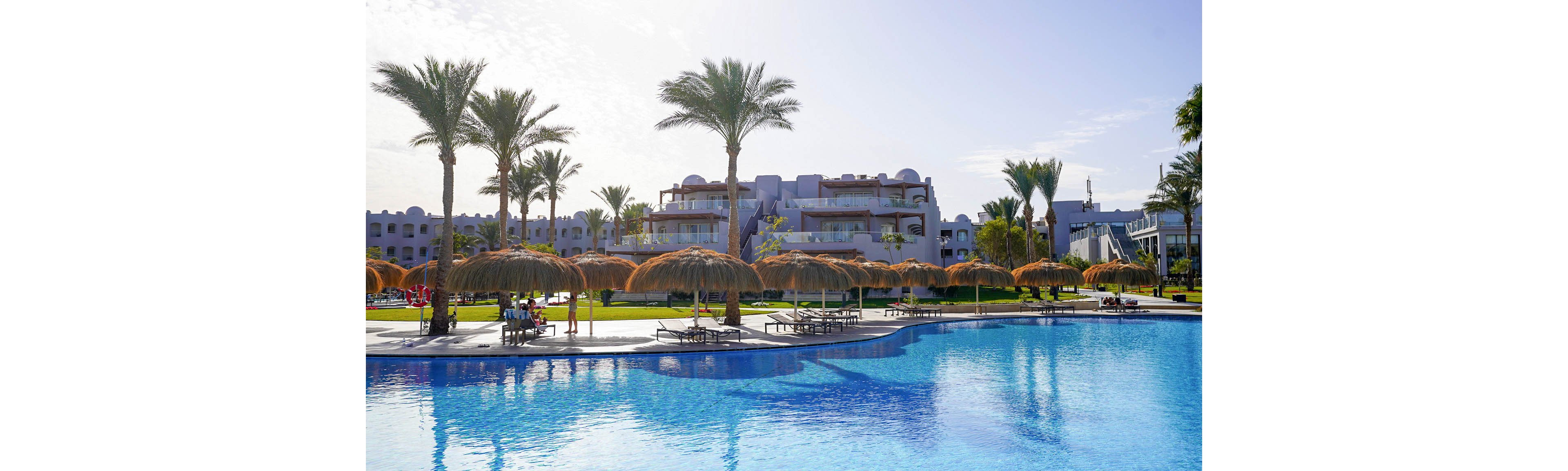 A large outdoor swimming pool reflects the clear blue sky in front of a modern resort complex. Tall palm trees and thatched sun umbrellas line the poolside, creating a tropical vacation atmosphere. Lounge chairs are arranged neatly under the umbrellas, with a few people relaxing near the water. The buildings in the background feature balconies and soft, neutral colors, adding to the calm and inviting setting.