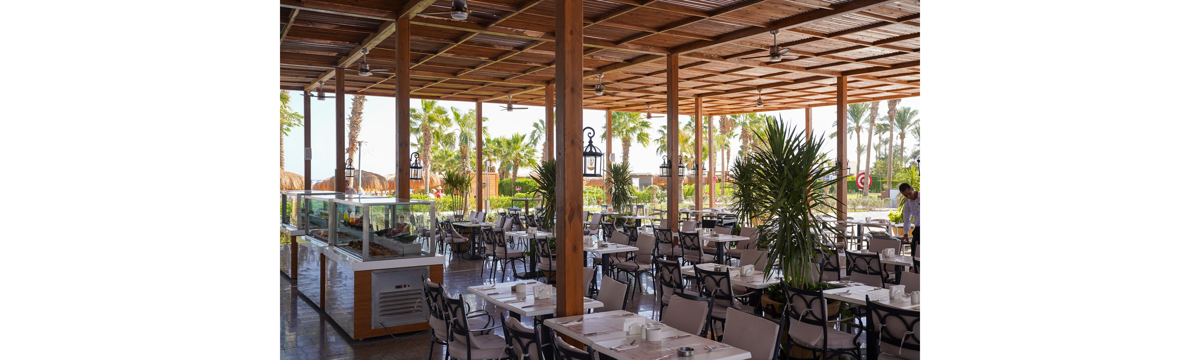 An open-air restaurant features rows of empty tables and chairs neatly arranged under a wooden pergola roof. White tableware is set on each table, and a glass buffet counter runs along one side of the space. Tall green plants and palm trees surround the dining area, suggesting a warm coastal or resort setting. A person stands near the edge of the terrace, with bright daylight illuminating the relaxed, vacation-like atmosphere.