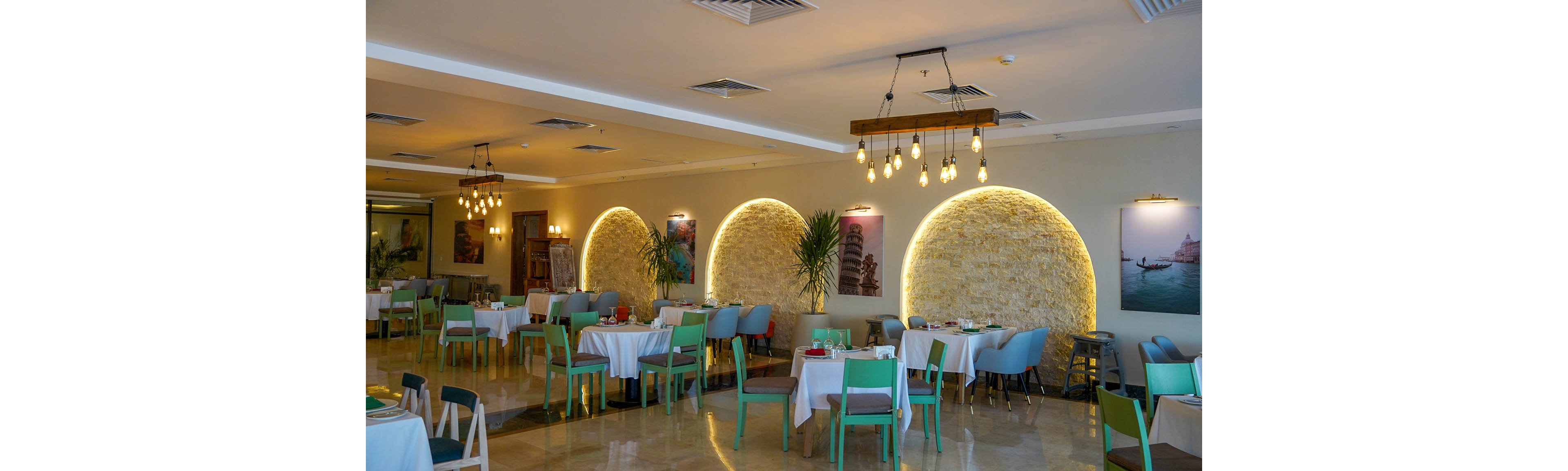 Spacious restaurant dining room with neatly arranged tables and chairs. The setting features contemporary decor, including arched stone accent walls with warm backlighting and hanging pendant lights. Tables are covered with white tablecloths and set for service, while colorful seating adds contrast to the neutral walls and floor. Framed wall art and indoor plants contribute to a relaxed, inviting atmosphere.