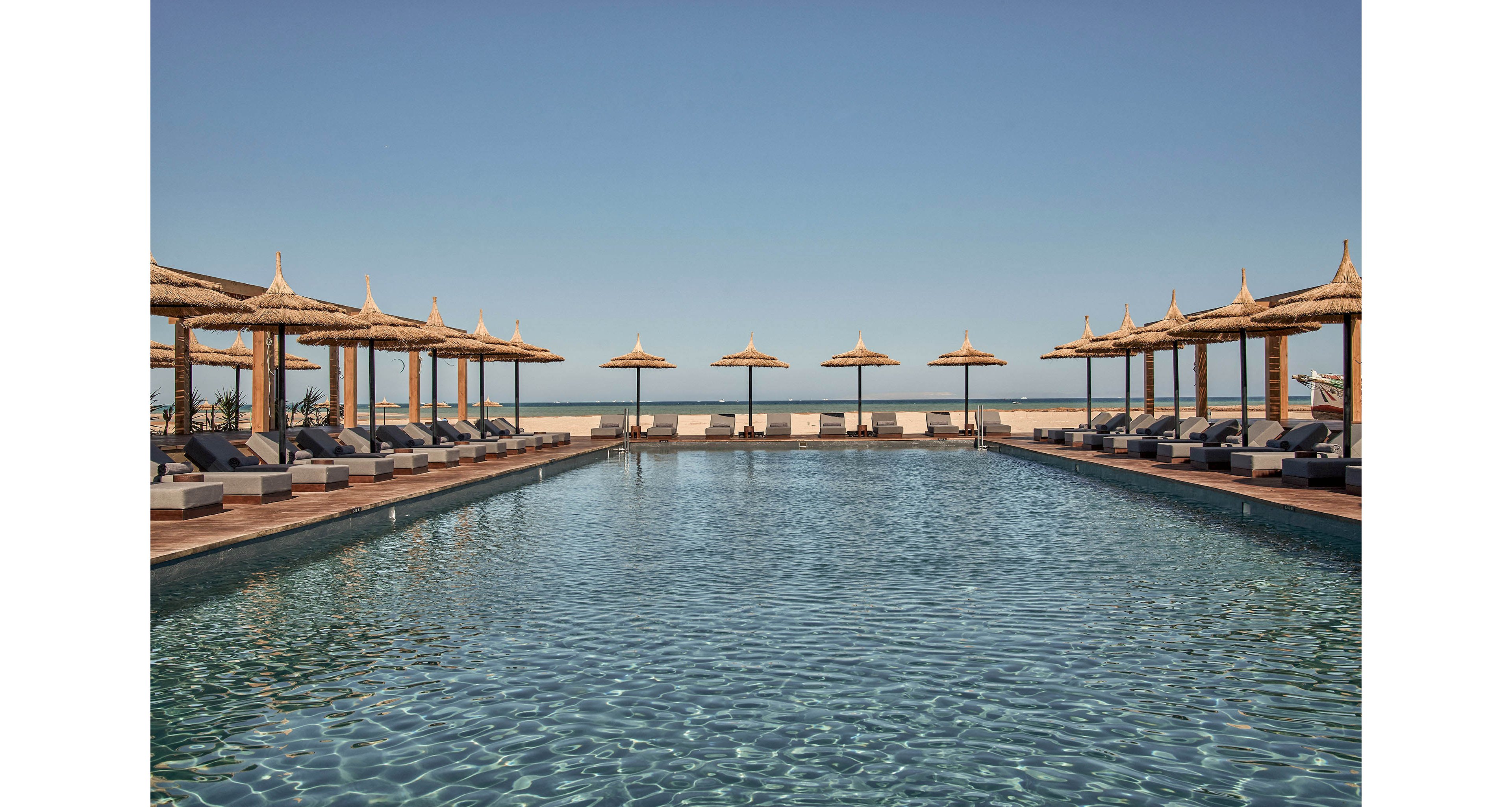 A serene outdoor pool surrounded by straw umbrellas and lounge chairs under a clear blue sky. The tranquil setting overlooks a distant horizon, creating a relaxing atmosphere. The water reflects the sunlight, enhancing the peaceful ambiance.