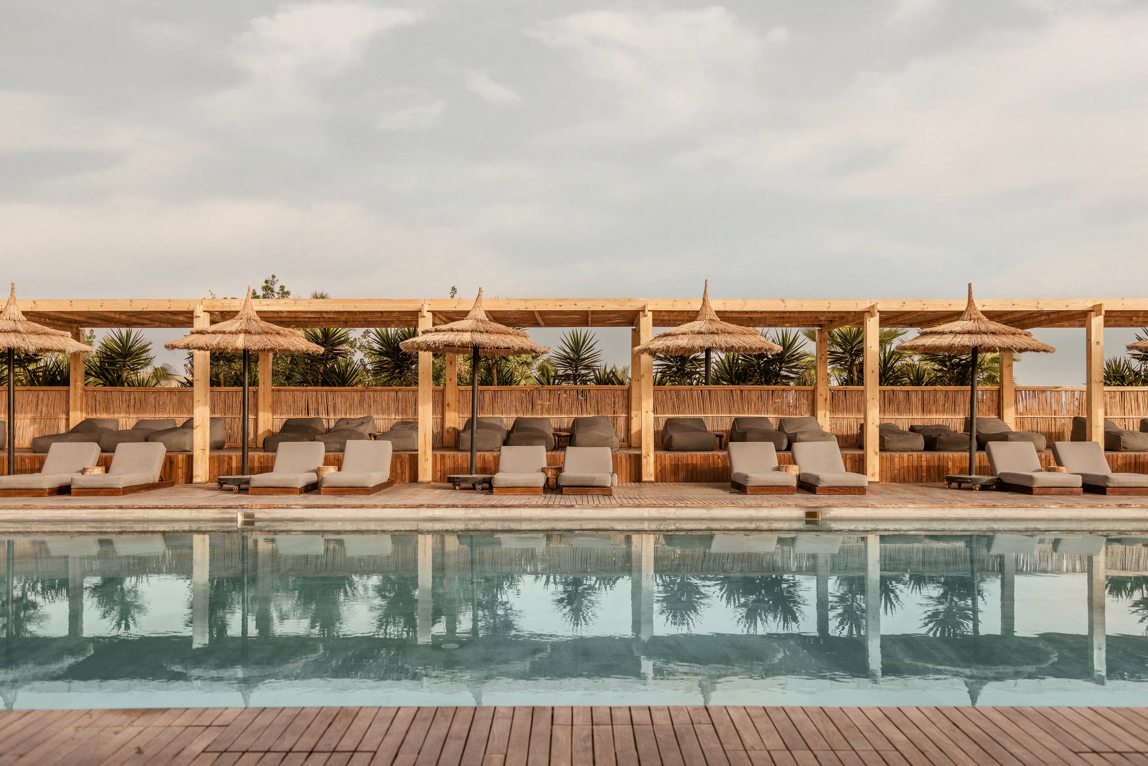 A serene outdoor pool surrounded by wooden loungers and straw umbrellas, set against a backdrop of lush greenery. Two people are seen enjoying the tranquil atmosphere, one seated and one walking by the poolside. The setting evokes relaxation and leisure under a partly cloudy sky.