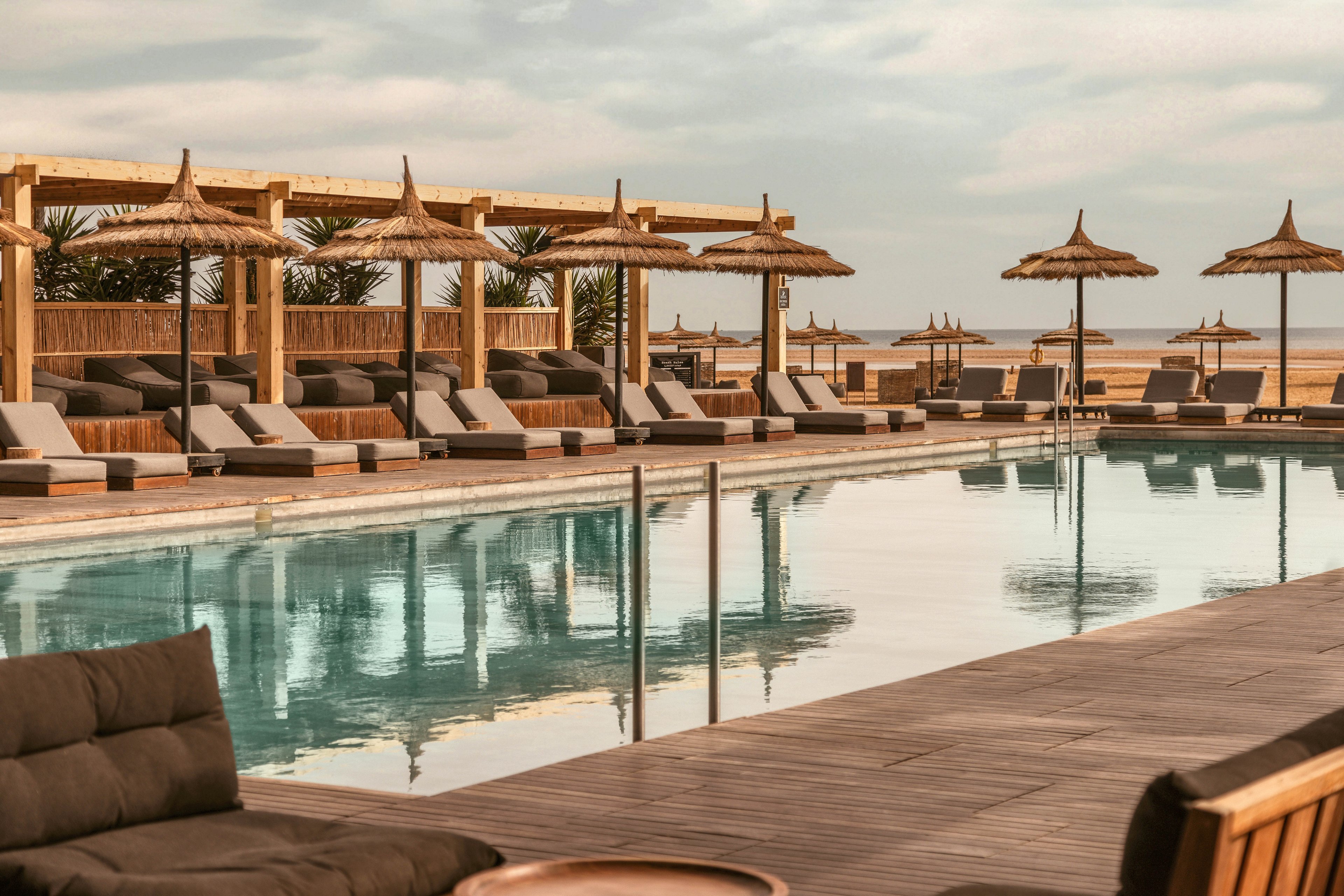 A serene outdoor pool surrounded by wooden lounge chairs and straw parasols, set against a backdrop of the ocean. The setting exudes a tranquil and luxurious vibe, perfect for relaxation. The warm tones and natural materials create a harmonious and inviting atmosphere.