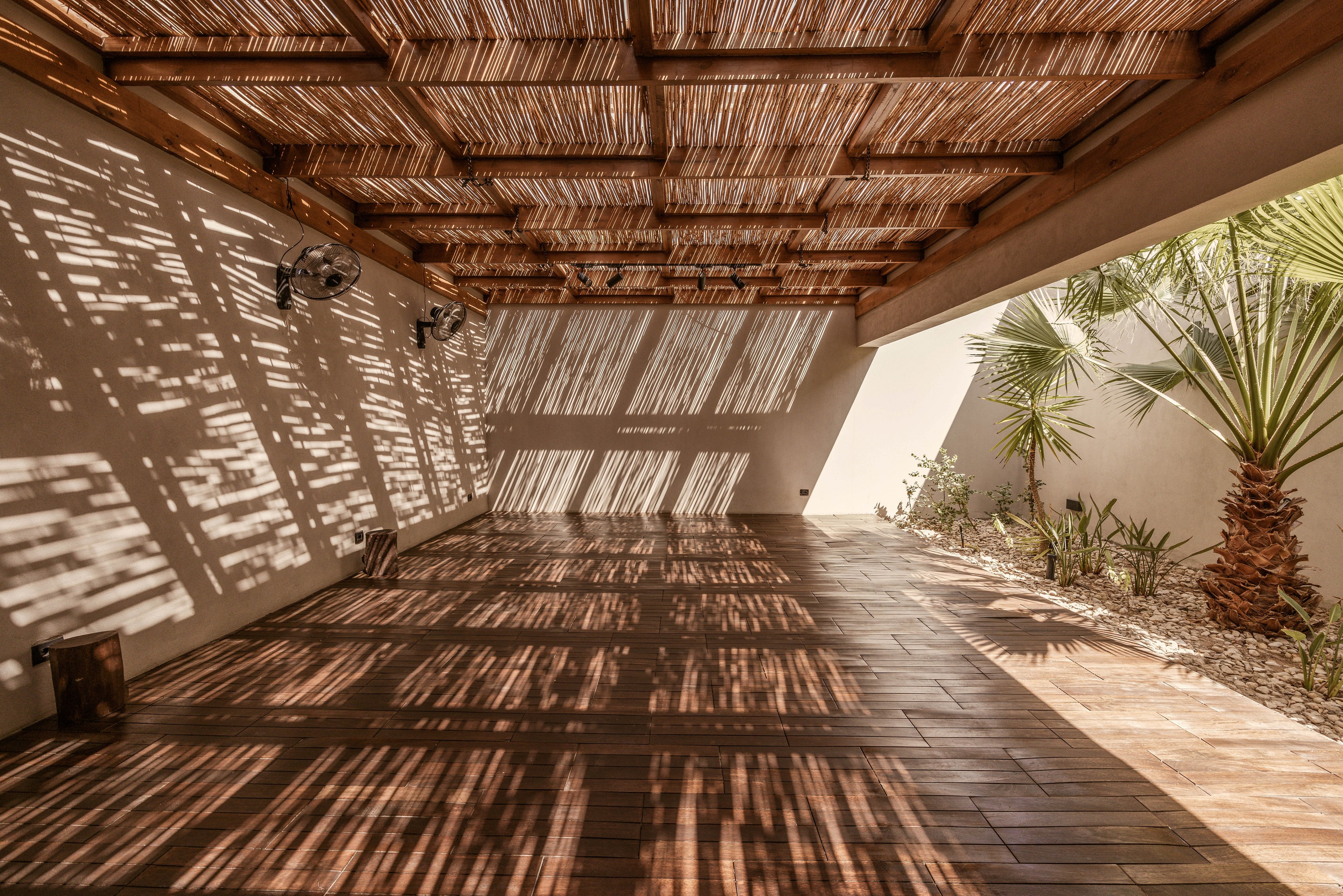 A spacious outdoor patio featuring a wooden pergola casting intricate shadows on the tiled floor. The setting includes tropical plants and a bright, sunny ambiance. The natural tones and textures create a warm and inviting atmosphere, ideal for relaxation or gatherings.