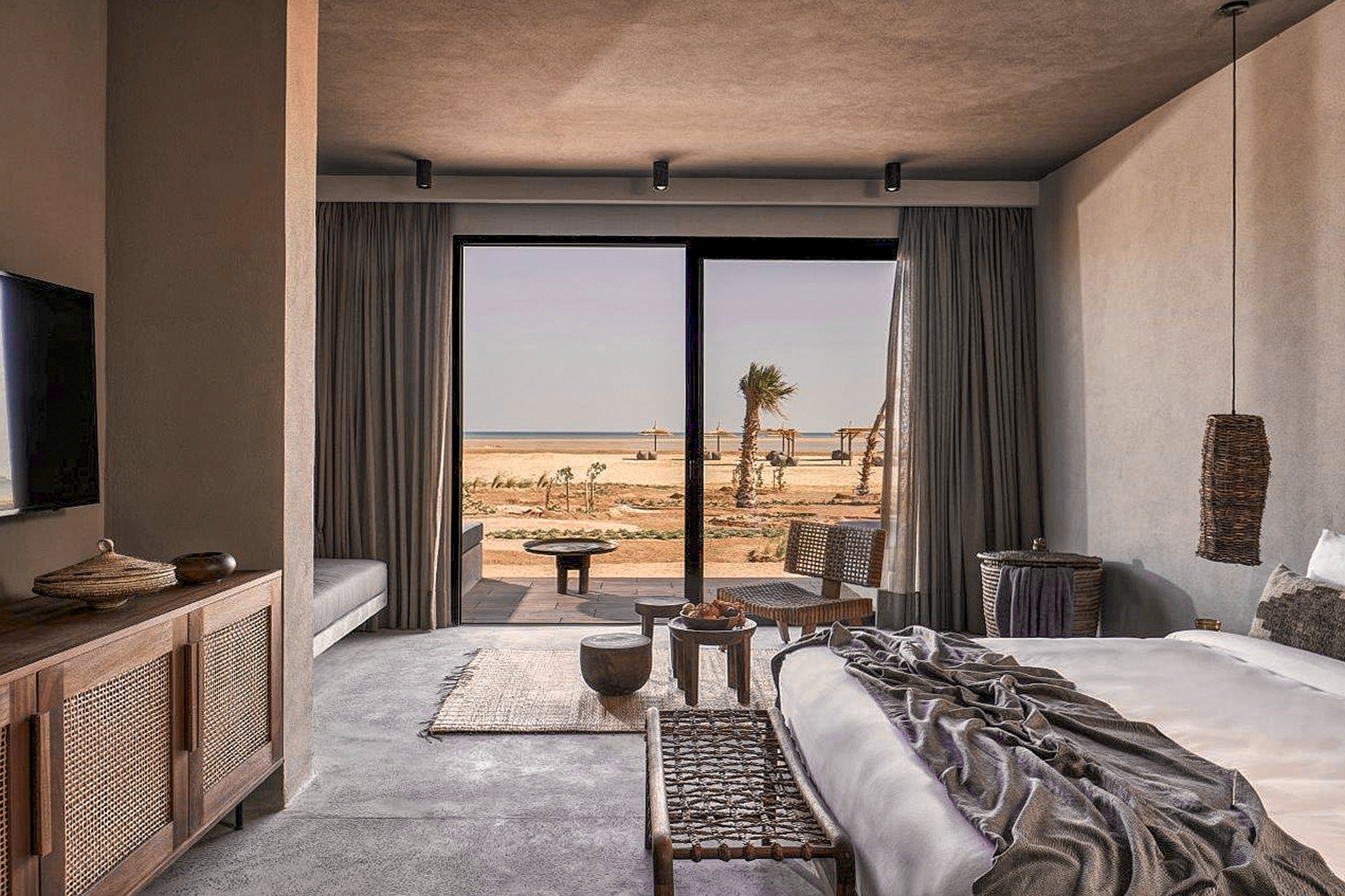 A stylish and modern hotel room featuring a cozy bed, woven furniture, and neutral tones. The room opens to a large glass door showcasing a serene beachfront view with palm trees and sandy terrain. The decor includes natural textures and minimalist lighting, creating a relaxing atmosphere.
