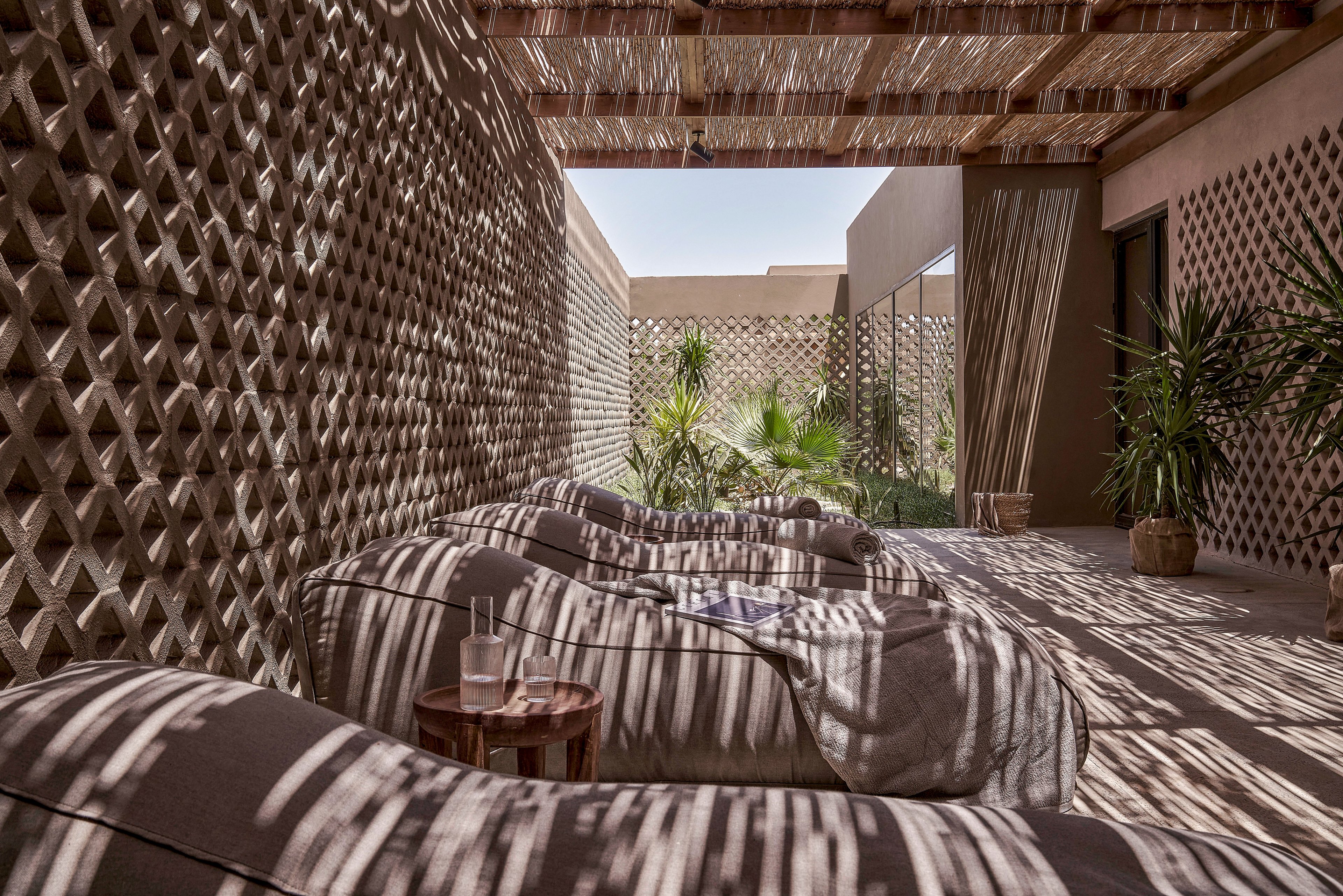 A serene outdoor lounge area featuring cushioned seating under a woven pergola that casts intricate shadow patterns. The setting includes natural tones, potted plants, and a small wooden side table with glasses. The space exudes a tranquil and earthy ambiance, perfect for relaxation.