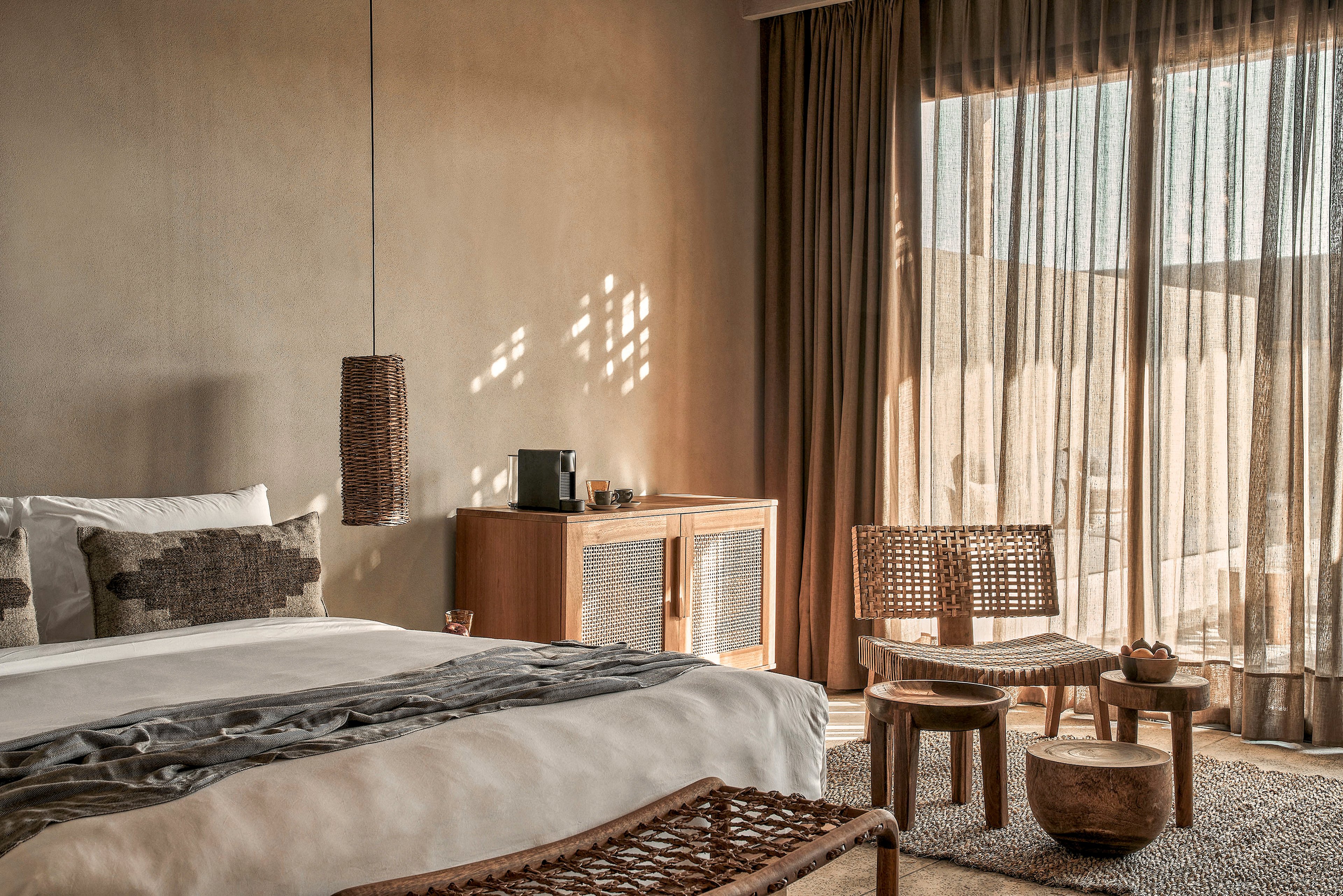 A serene bedroom featuring a large bed with neutral-toned bedding, wicker furniture, and wooden accents. The room is illuminated by natural light streaming through sheer curtains, creating a warm and inviting atmosphere. A small cabinet and pendant light add to the minimalist, earthy aesthetic.