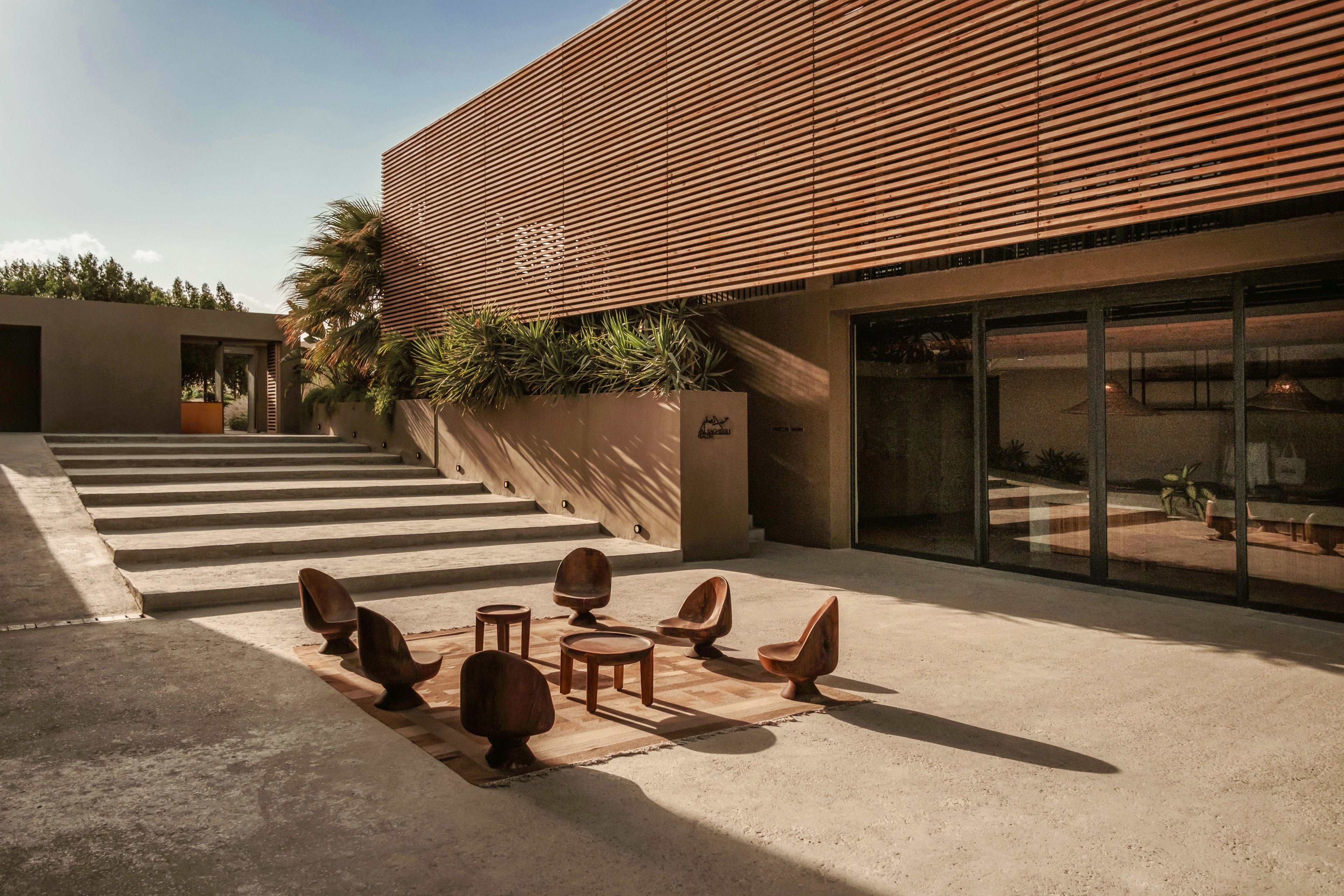 A contemporary courtyard featuring a minimalist seating arrangement with six chairs and a central table. The setting is surrounded by wooden paneling and concrete steps, complemented by greenery and soft lighting. The warm, earthy tones create a serene and inviting atmosphere.