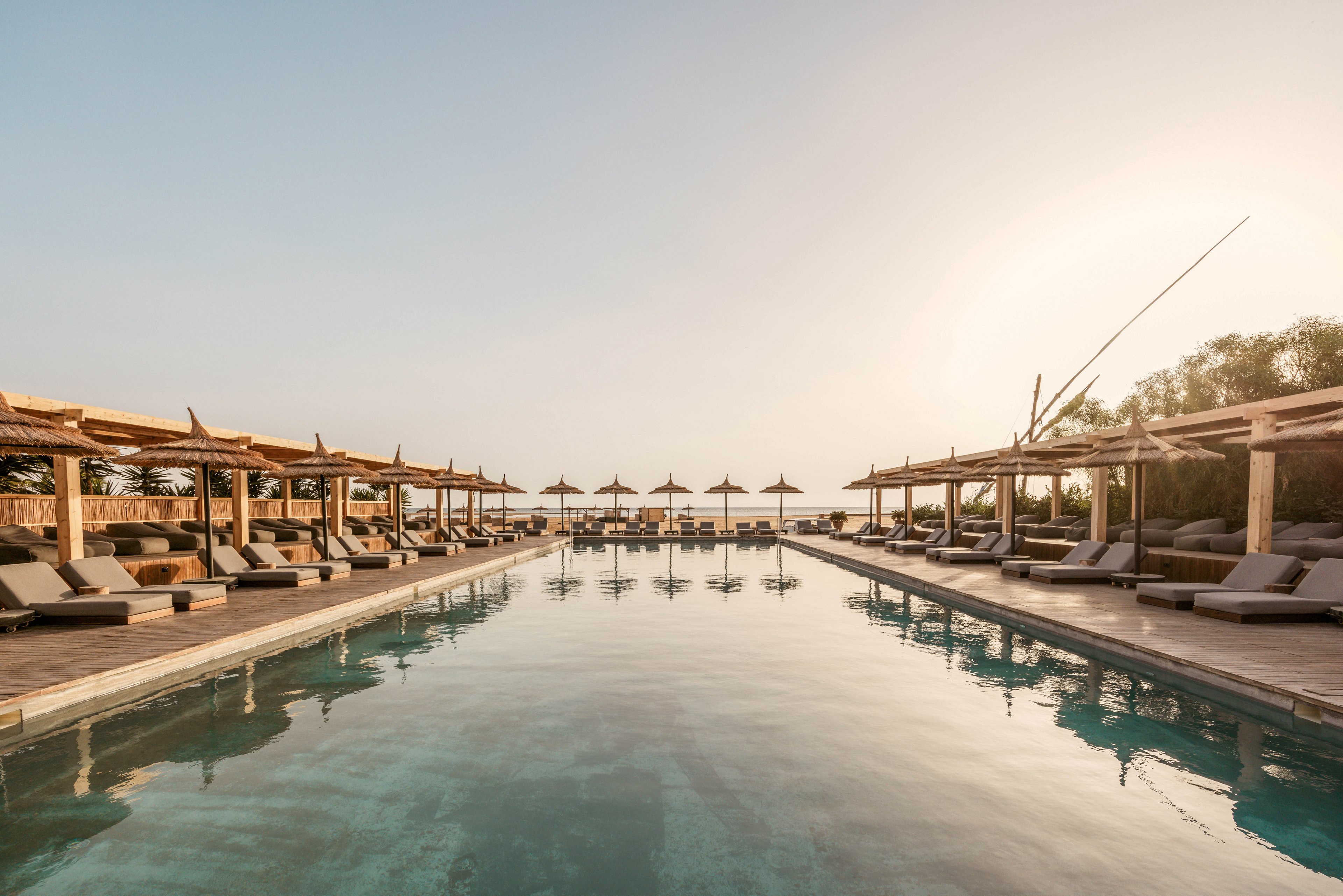 A serene outdoor pool surrounded by wooden sun loungers and umbrellas, set in a luxurious resort. The setting features a tranquil atmosphere with warm lighting during sunset. The symmetrical design and calm water create a relaxing and inviting ambiance.