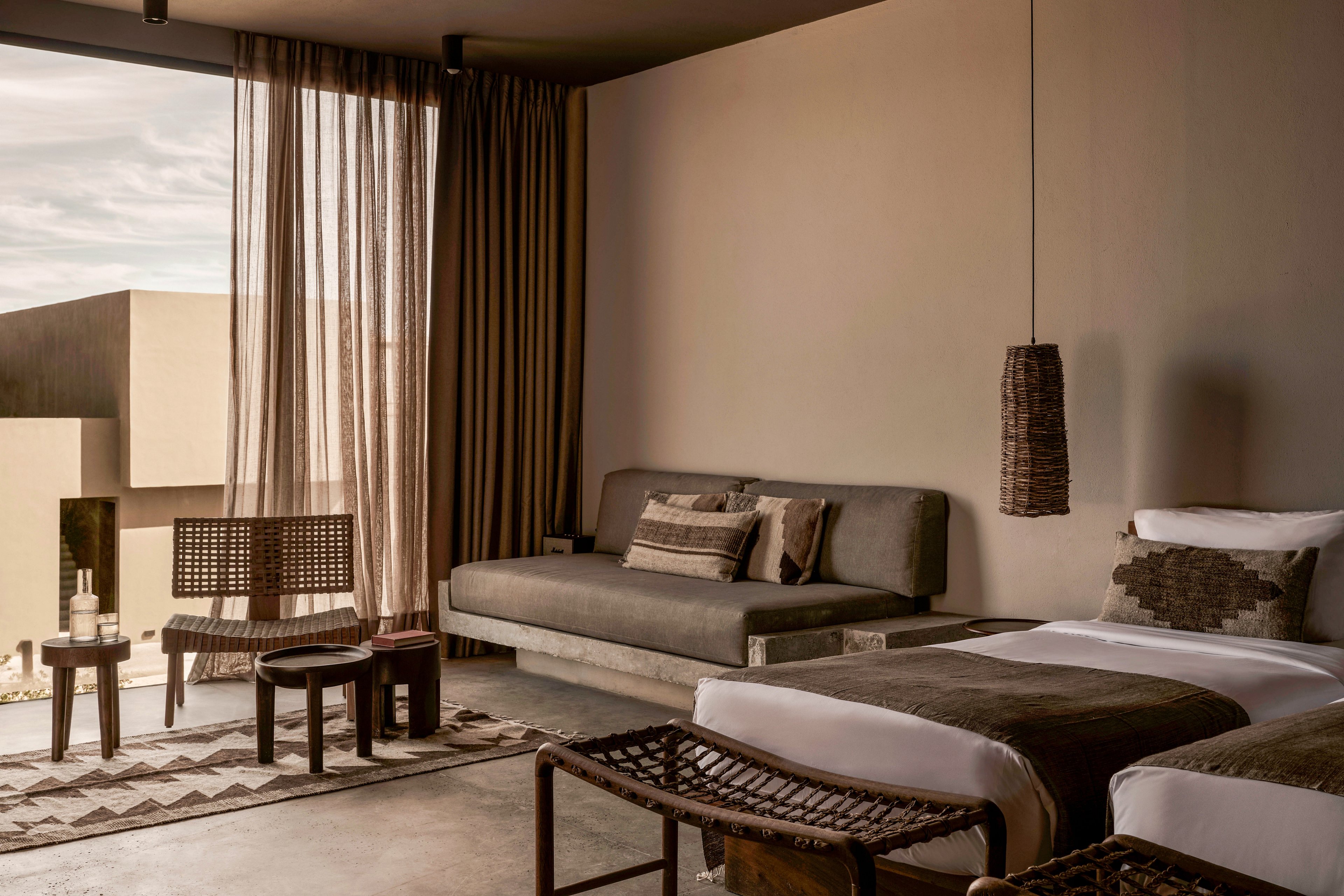 A contemporary hotel room featuring minimalist decor with earthy tones and natural materials. The room includes a bed, a sofa, woven chairs, and a small table, with soft lighting and sheer curtains framing a view of an outdoor space. The design emphasizes simplicity and a calming atmosphere.