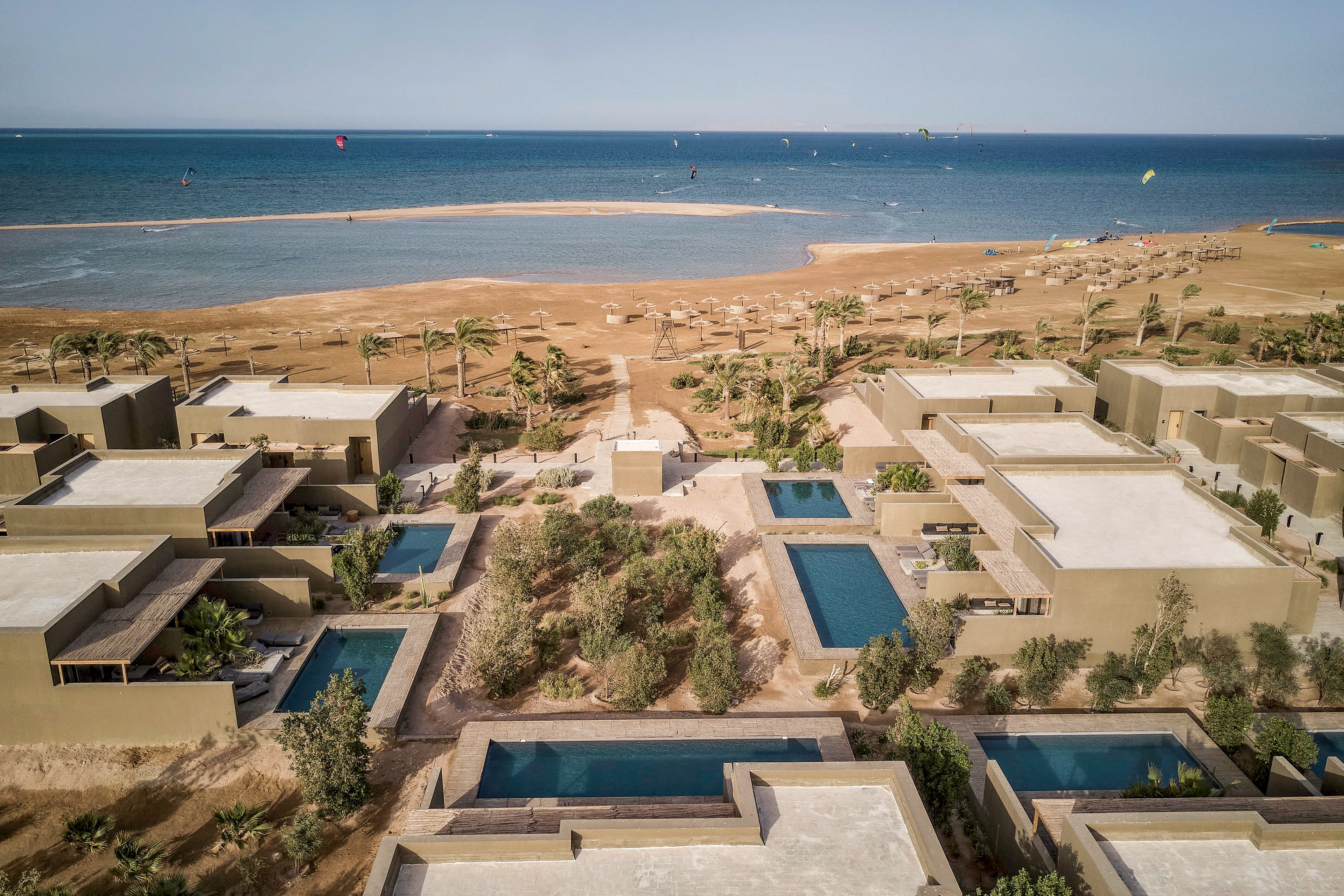 Aerial view of modern beachfront villas featuring private pools and lush landscaping. The setting overlooks a serene beach with calm waters and kite surfers in the distance. The scene captures a tranquil and luxurious coastal lifestyle under a clear blue sky.