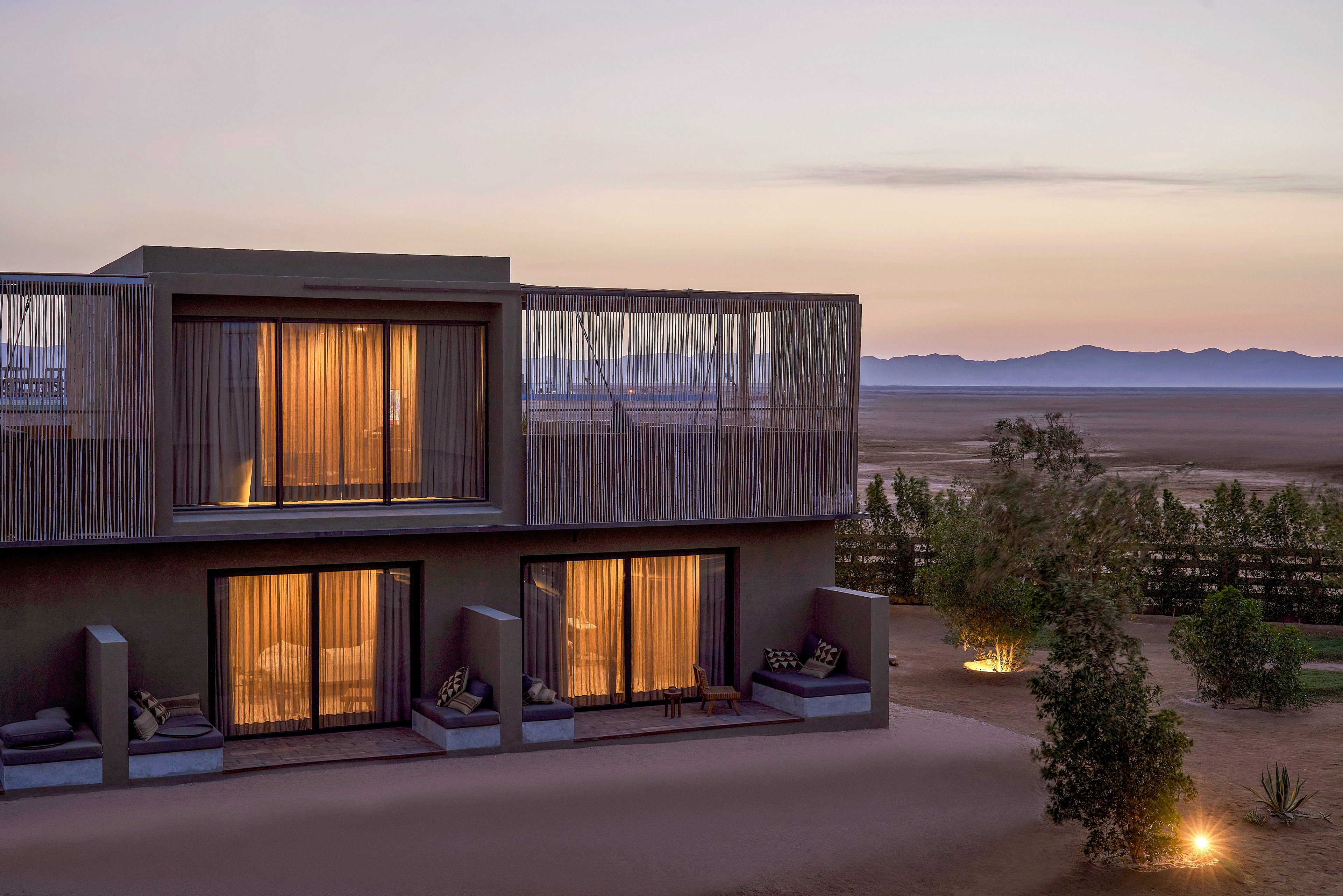 A contemporary two-story villa with illuminated interiors is set against a serene desert landscape at dusk. The building features large glass windows and a minimalist design, surrounded by sparse greenery and soft outdoor lighting. The distant mountains and pastel sky enhance the tranquil ambiance.