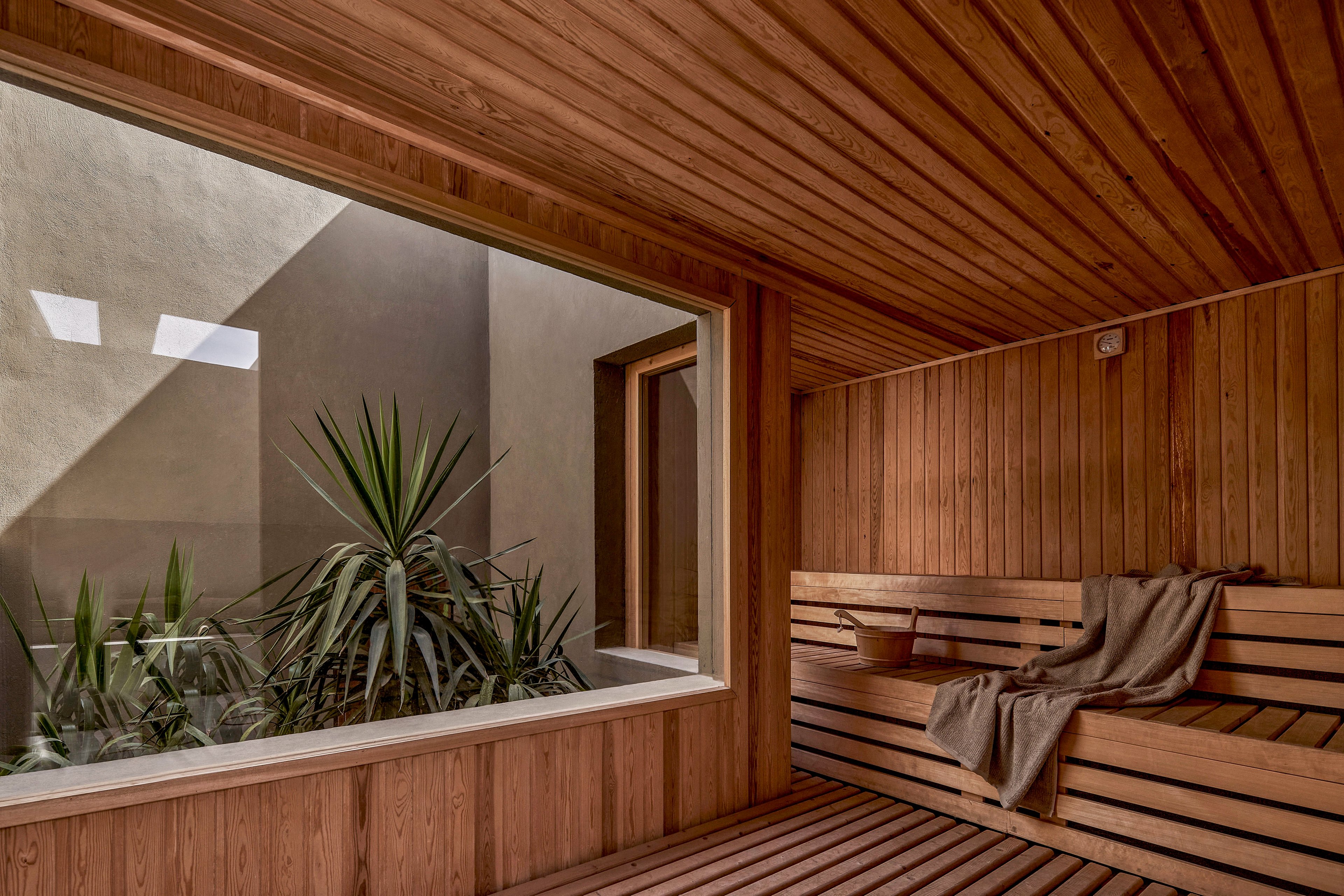 A contemporary sauna interior featuring wooden paneling and a large window overlooking a small garden with green plants. The space is illuminated by natural light streaming through the window, creating a serene and relaxing atmosphere. A towel is neatly placed on the wooden bench, adding to the inviting ambiance.