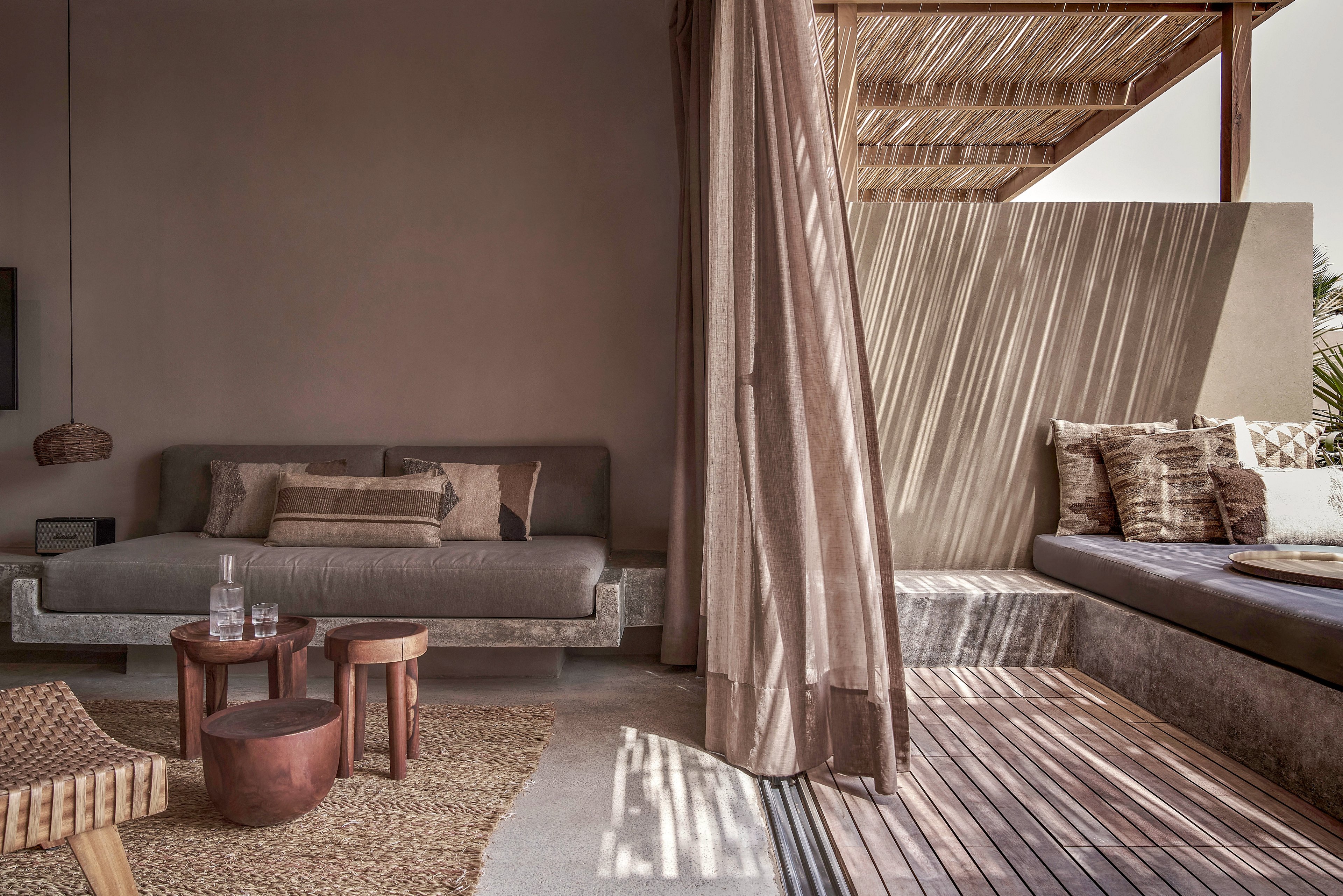 A stylish outdoor lounge area featuring a neutral color palette with a mix of beige, brown, and gray tones. The space includes a comfortable sofa, wooden chairs, and a small round table, complemented by natural light streaming through a woven canopy. The setting exudes a cozy and serene atmosphere, perfect for relaxation.