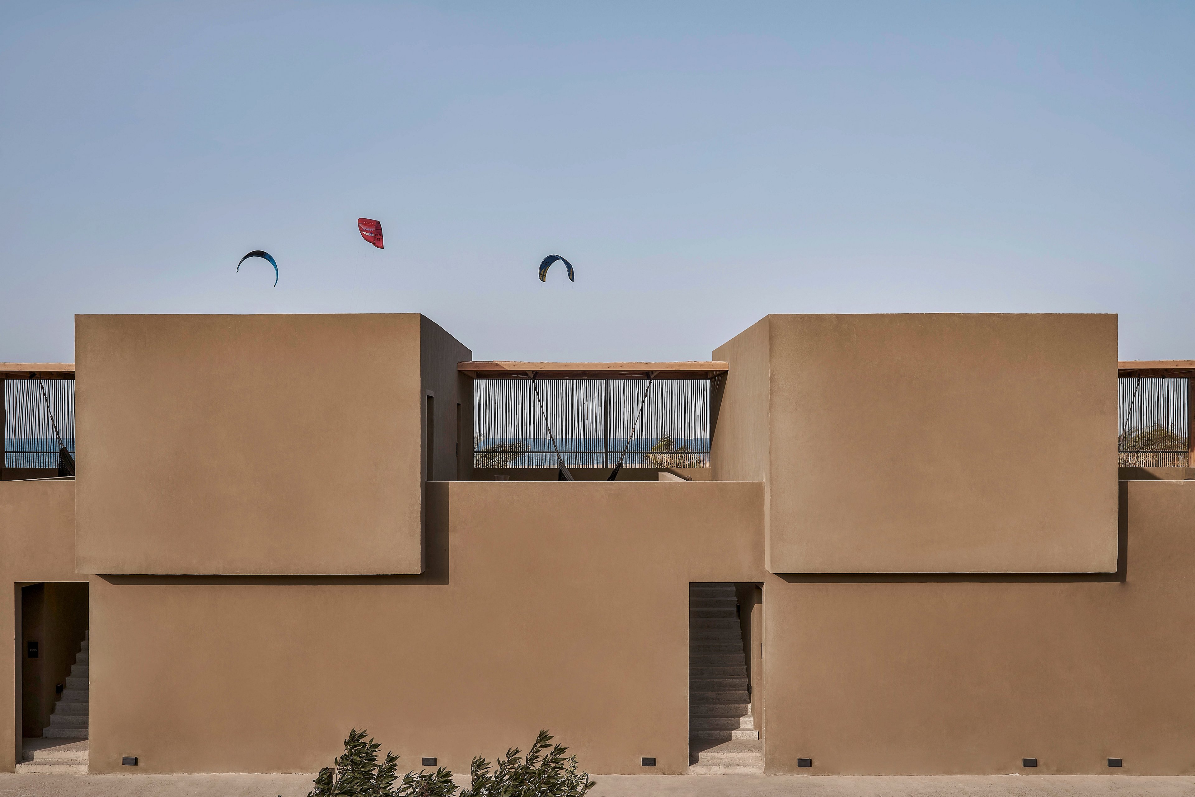 A minimalist building with geometric shapes stands in a desert-like setting under a clear blue sky. Kite surfers are visible in the background, adding a dynamic element to the serene environment. The muted beige tones of the structure contrast with the vibrant kites in the distance.