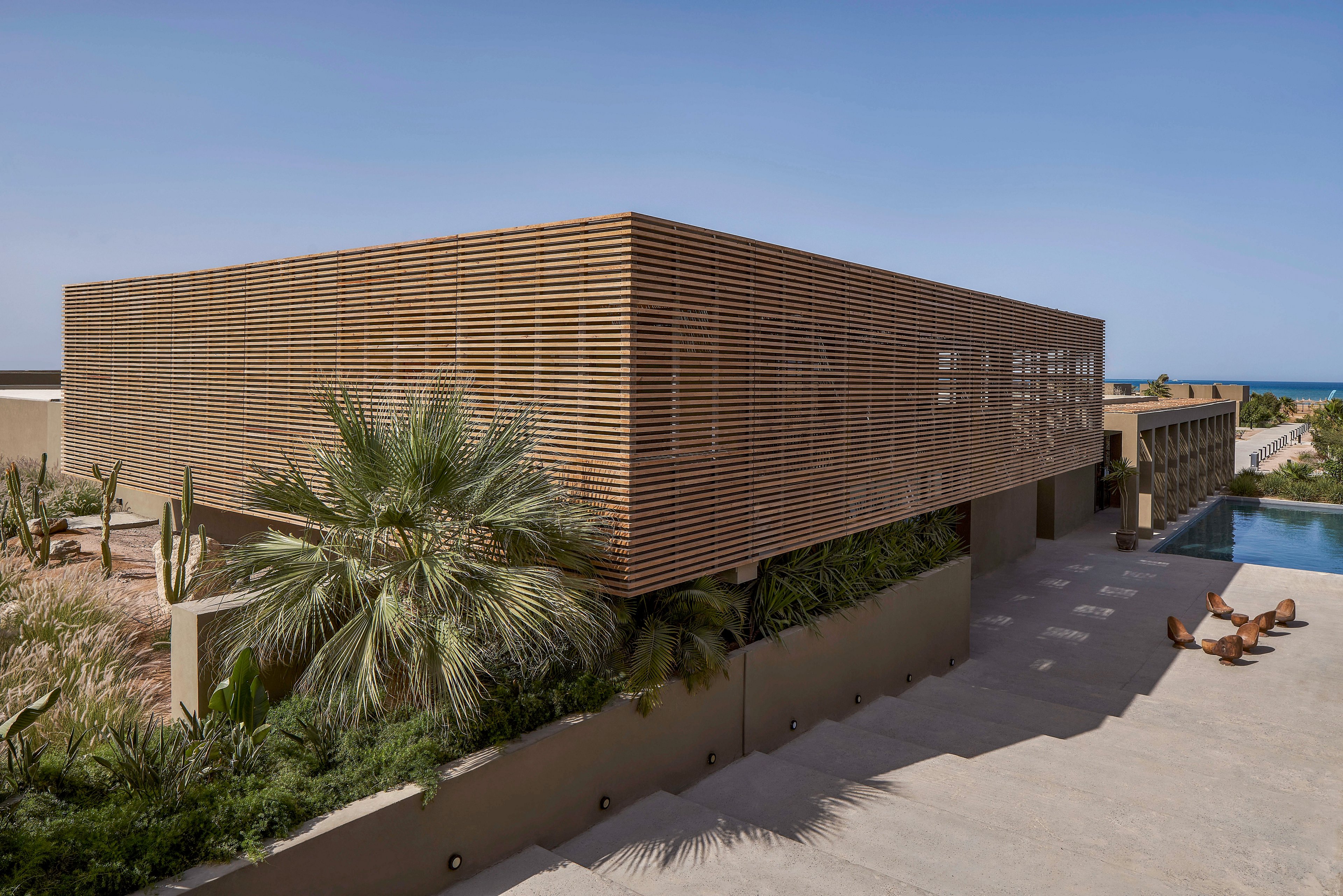 A contemporary building featuring a wooden slatted facade surrounded by lush greenery and desert plants. The setting includes a serene poolside area with lounge chairs under a clear blue sky. The minimalist architecture emphasizes clean lines and natural materials, creating a tranquil atmosphere.