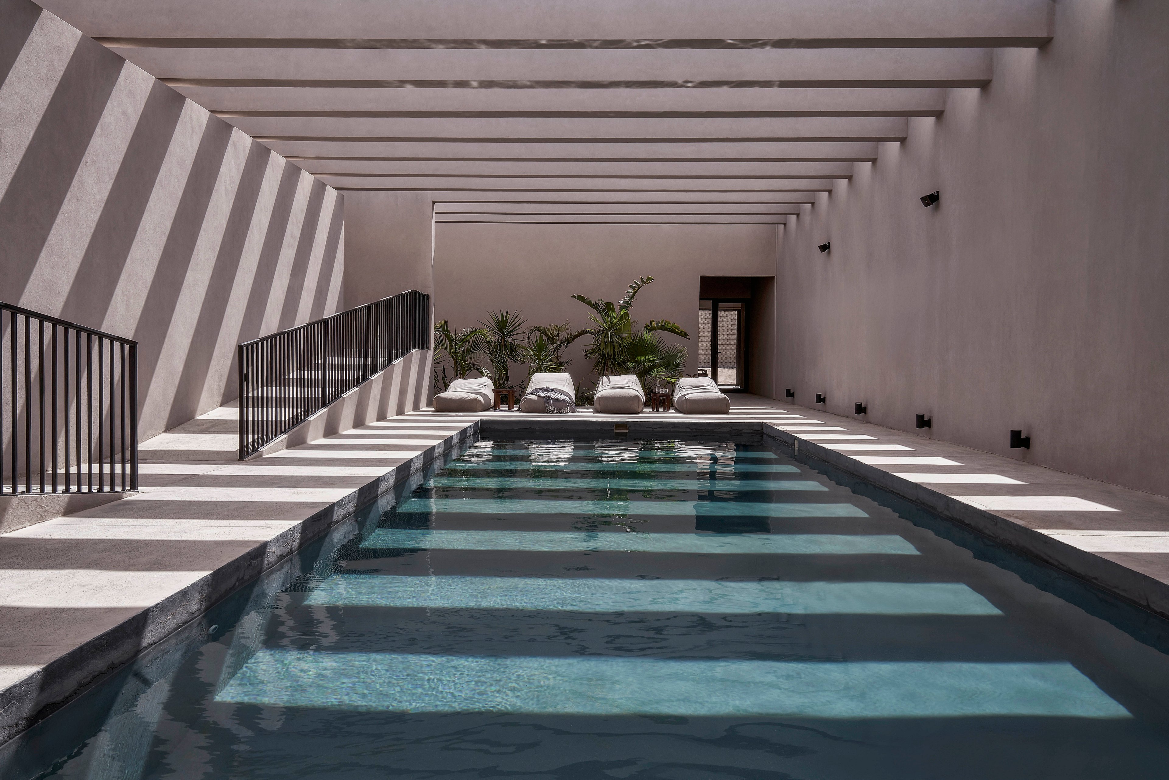 A sleek indoor pool with a minimalist design, featuring clean lines and a serene atmosphere. Four white lounge chairs are arranged at the far end, complemented by potted plants for a touch of greenery. The space is illuminated by natural light streaming through slatted beams, creating striking shadow patterns on the walls and water.